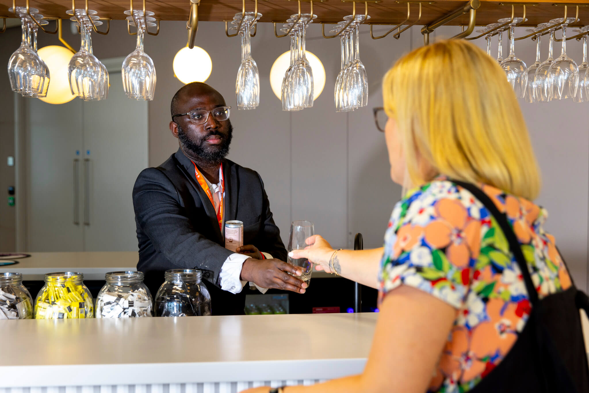 Male colleague serving female in floral top a drink across a bar. Glasses hanging from storage.