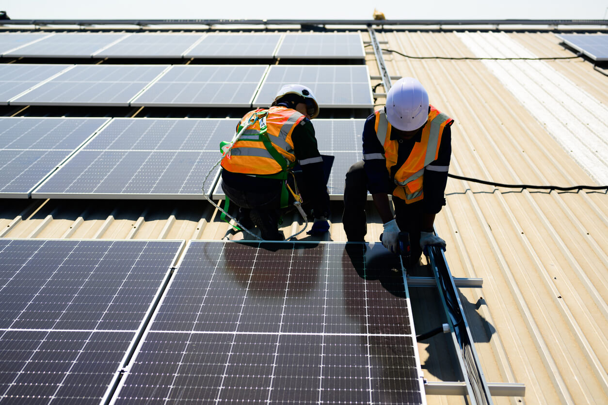 Two engineers wearing safety equipment. Both knelt down and installing solar panels to the roof. 