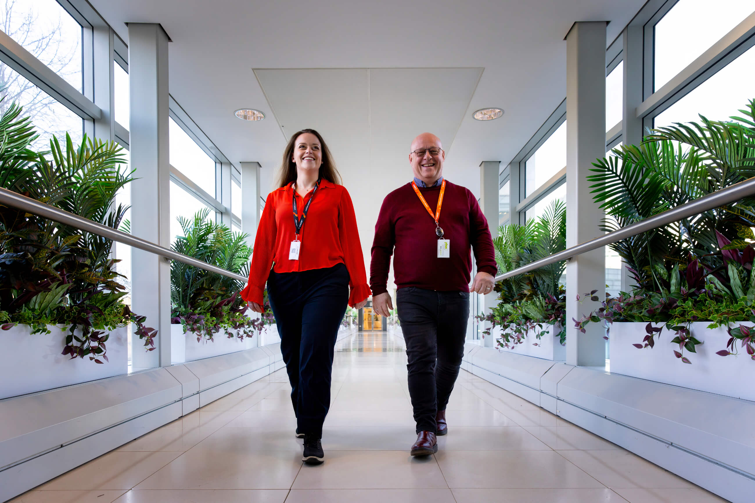 Two colleagues wearing office wear walking down a corridor. Plants and handrails either side of them. 