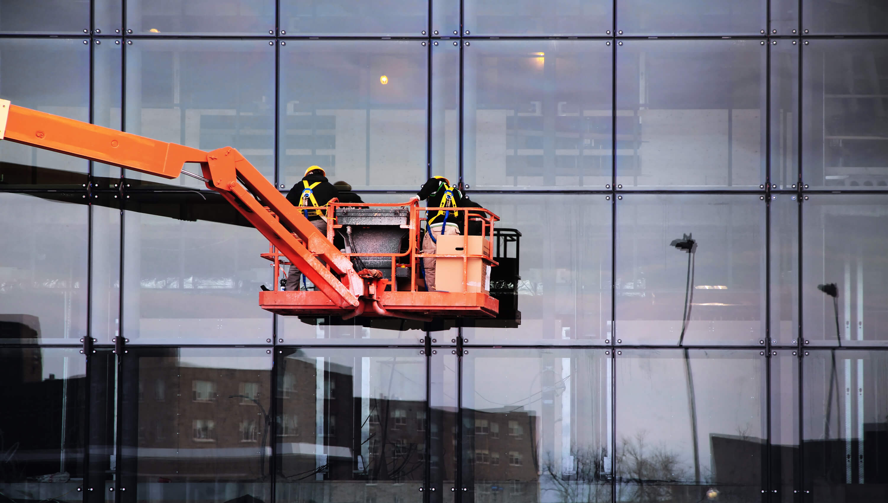 Two engineers on an orange cherry picker outside office building. Wearing harnesses and city reflecting in the windows.