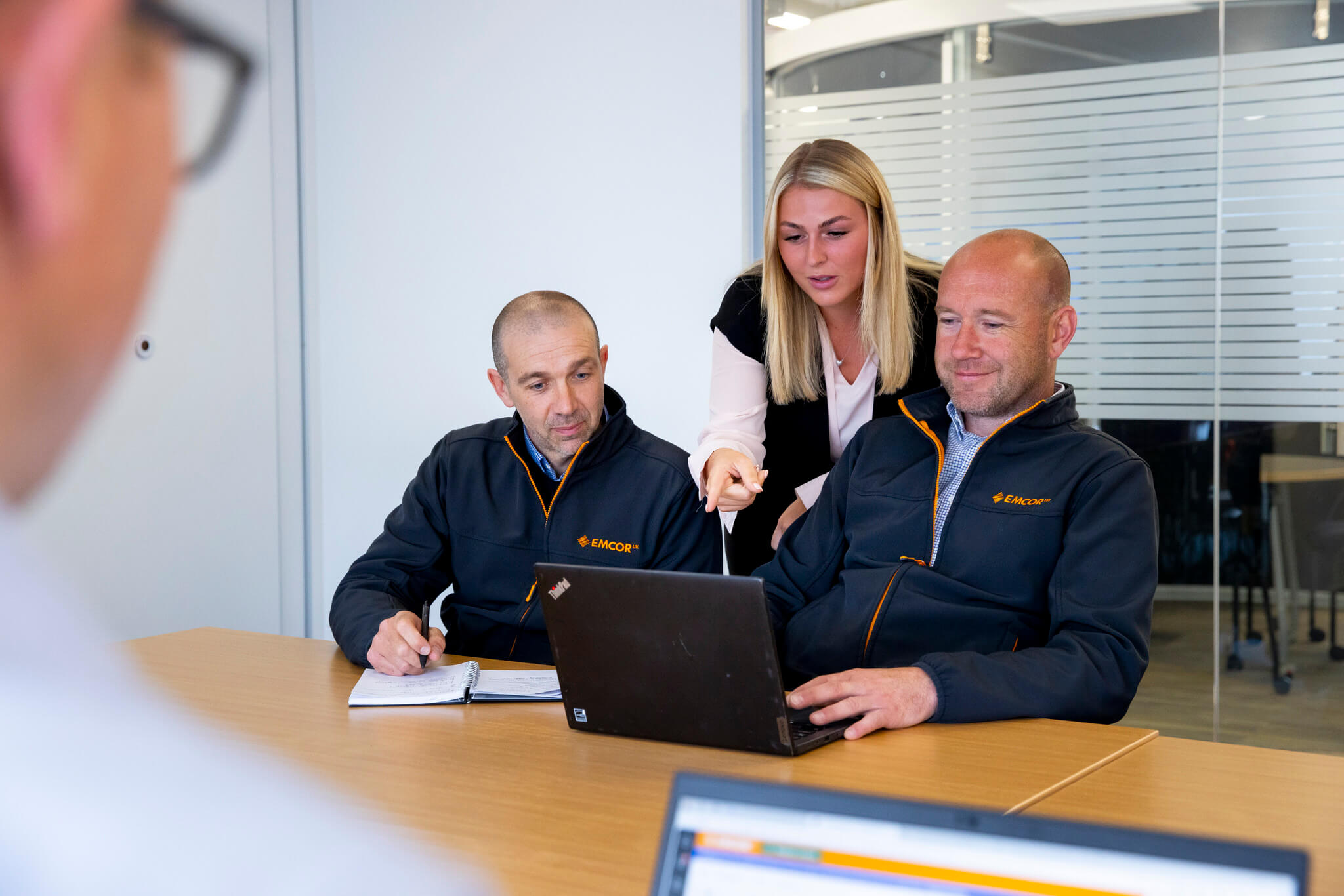 Female stood behind two male colleagues in a meeting room. Female is pointing at a laptop on the desk. 