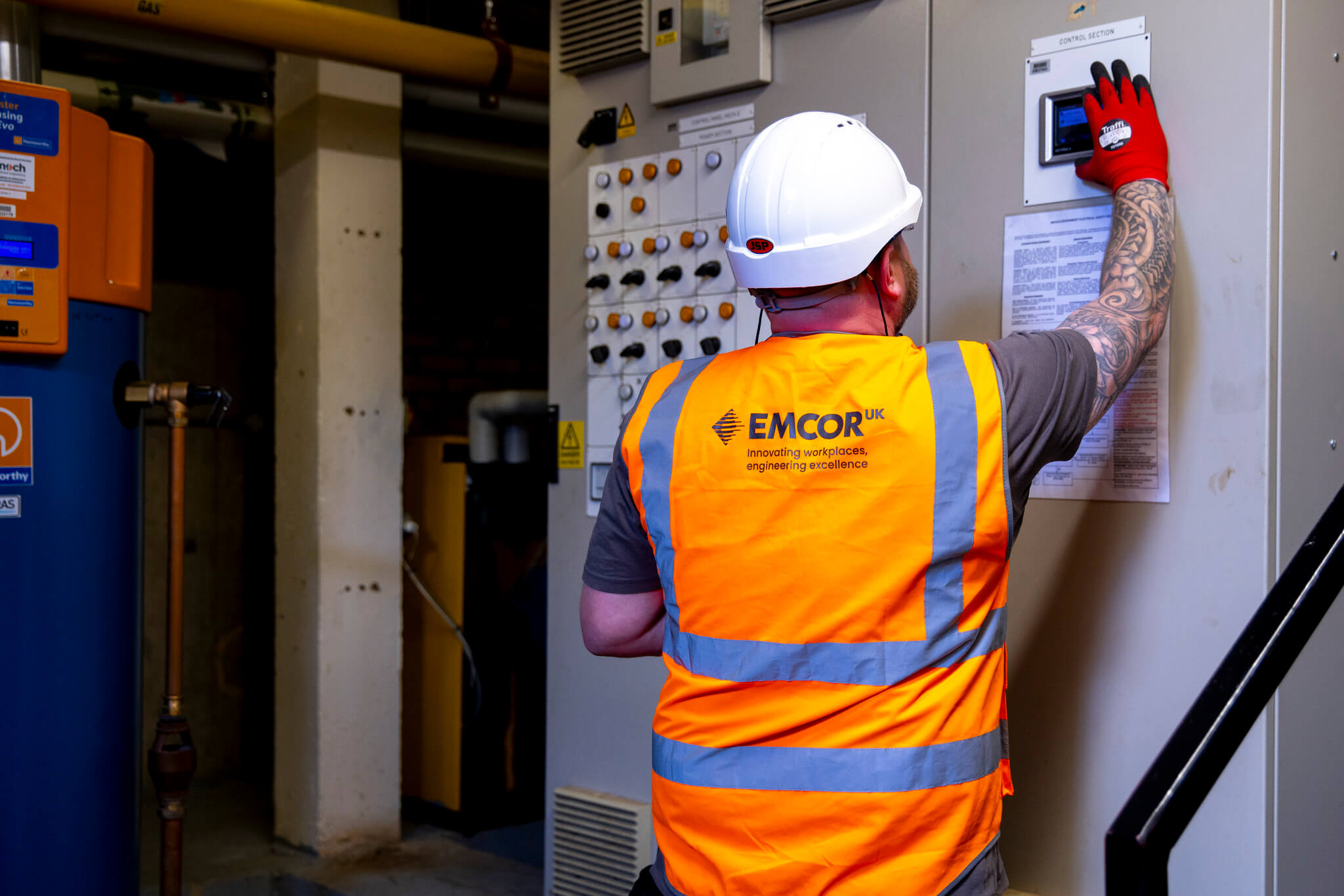 Male engineer wearing orange high vis and white hard hat with red gloves pressing buttons on a control panel.