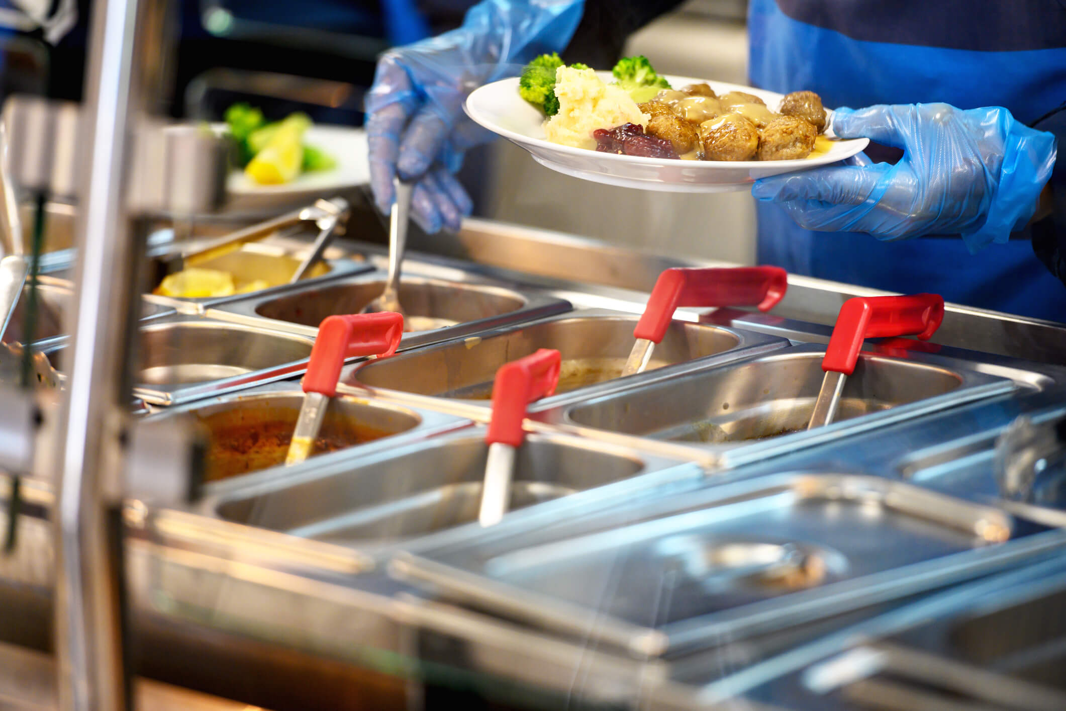 Person wearing blue gloves and apron holding ladle and plate of food. Containers of hot food in front of them. 