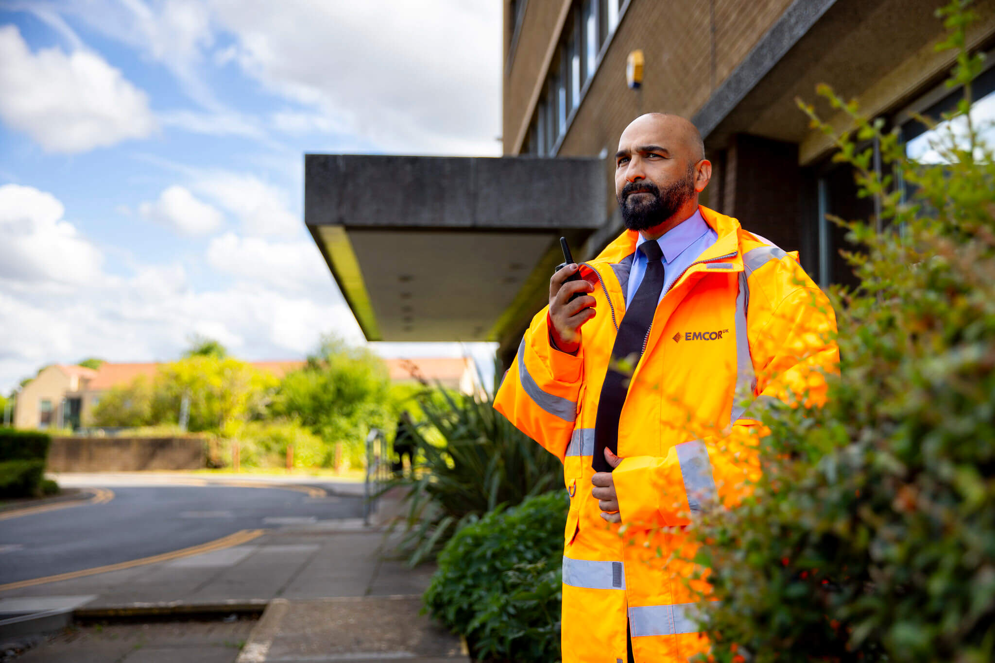 Man in an orange safety jacket holds a radio outside a building, with greenery and a street in the background. 