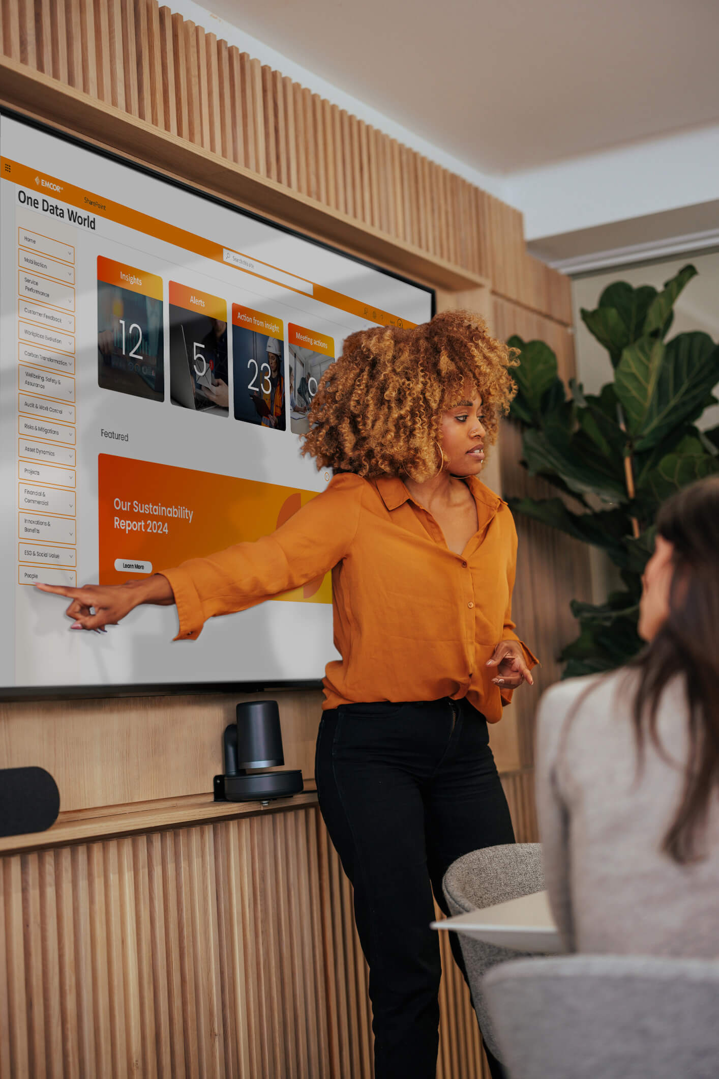 Female wearing orange shirt presenting on a large screen in modern meeting room.