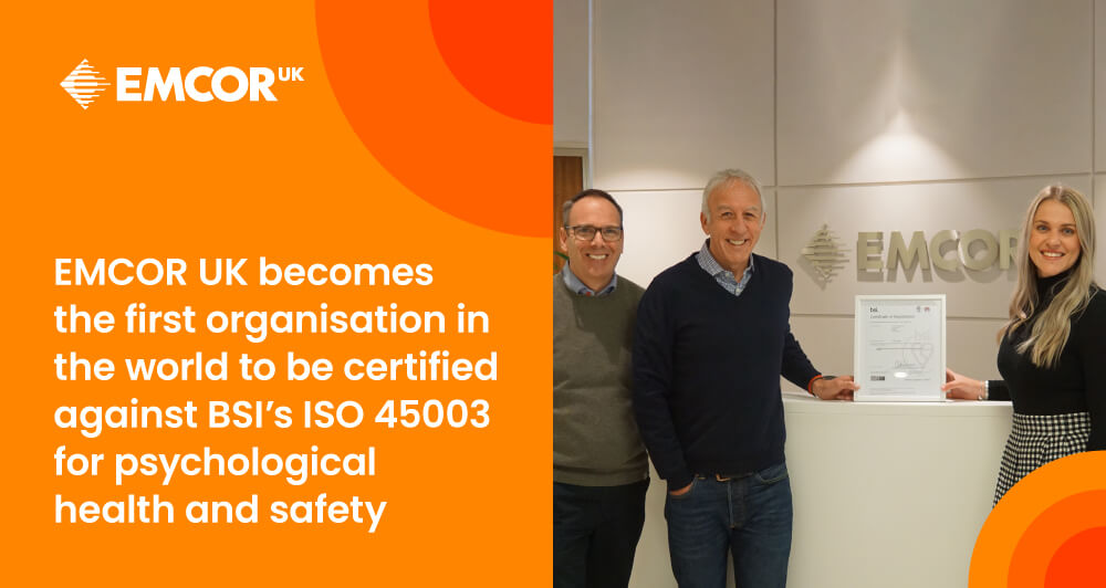 Three EMCOR UK executives standing in the lobby holding a plaque celebrating their certification for the BSI ISO 45003 program.