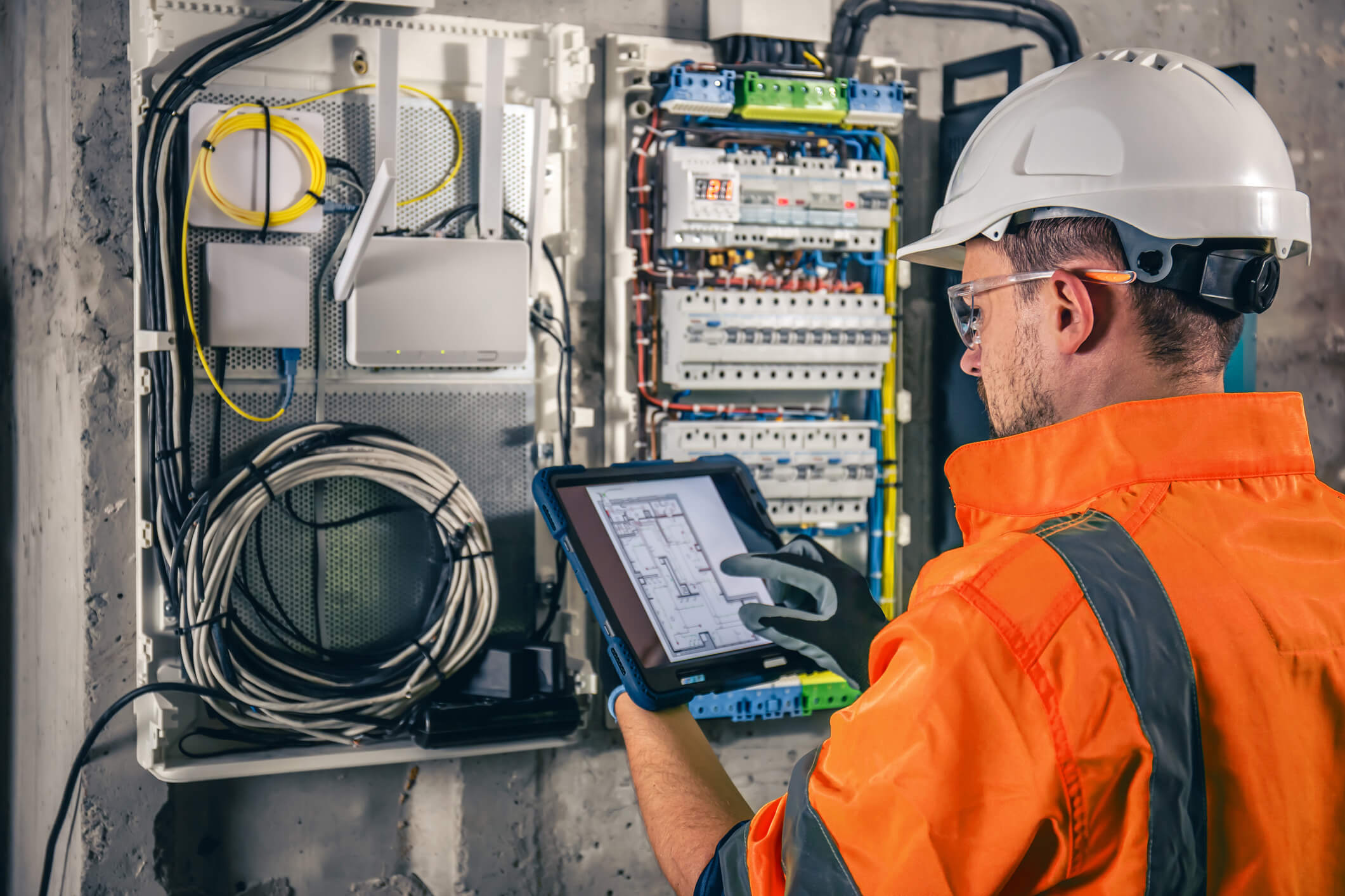 Male engineer wearing orange high vis, hard hat and glasses holding an iPad. Switchboard in the background with wires.