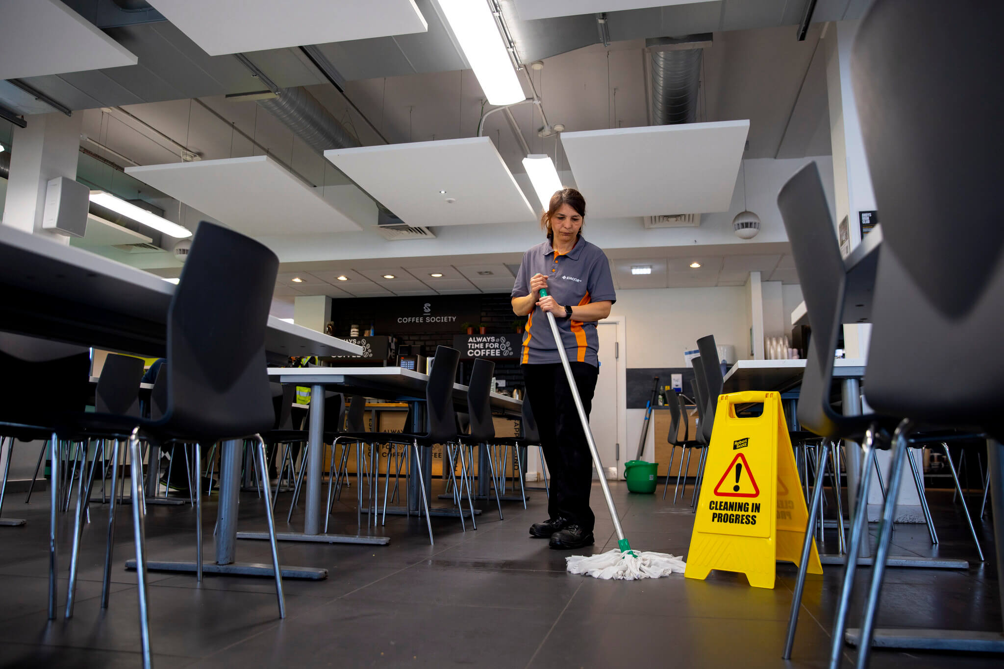 Female cleaner mopping floors of office canteen space. Cleaning in progress sign on the floor.