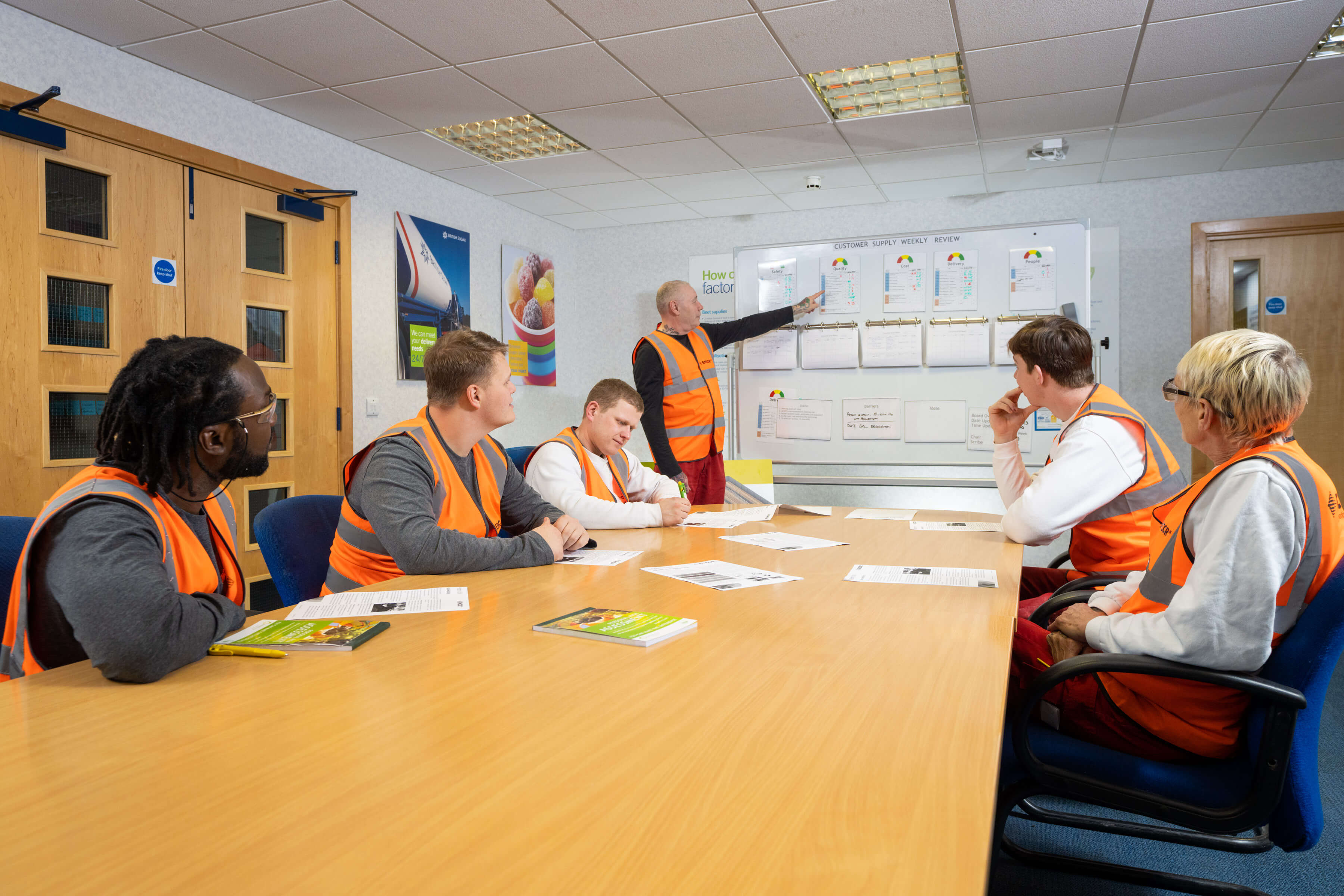 Male trainer pointing to whiteboard in front of a group of trainees sat at a table all wearing high-vis waistcoats.