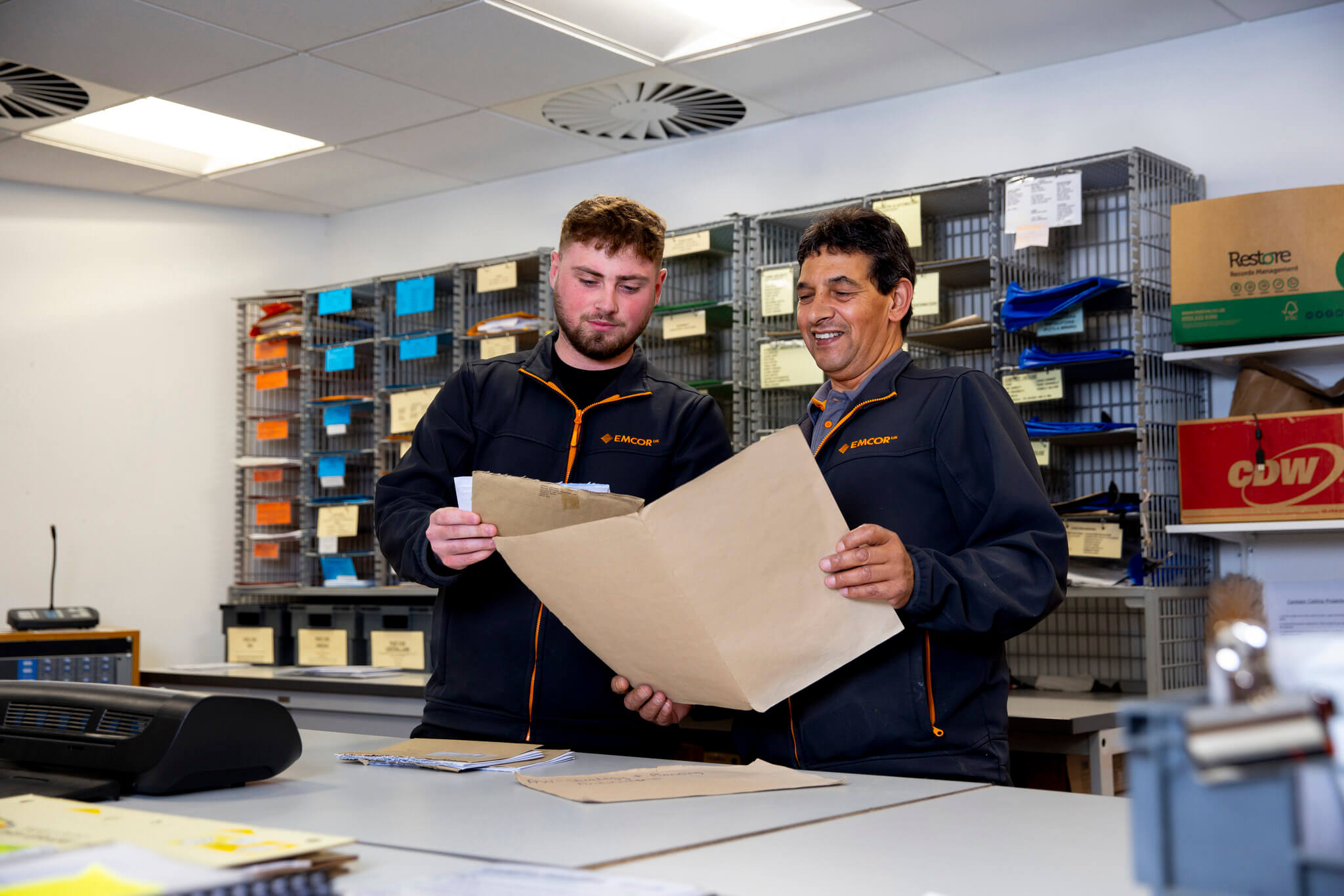 Two male colleagues wearing EMCOR UK uniform in post room holding files. Pigeonholes in the background. 