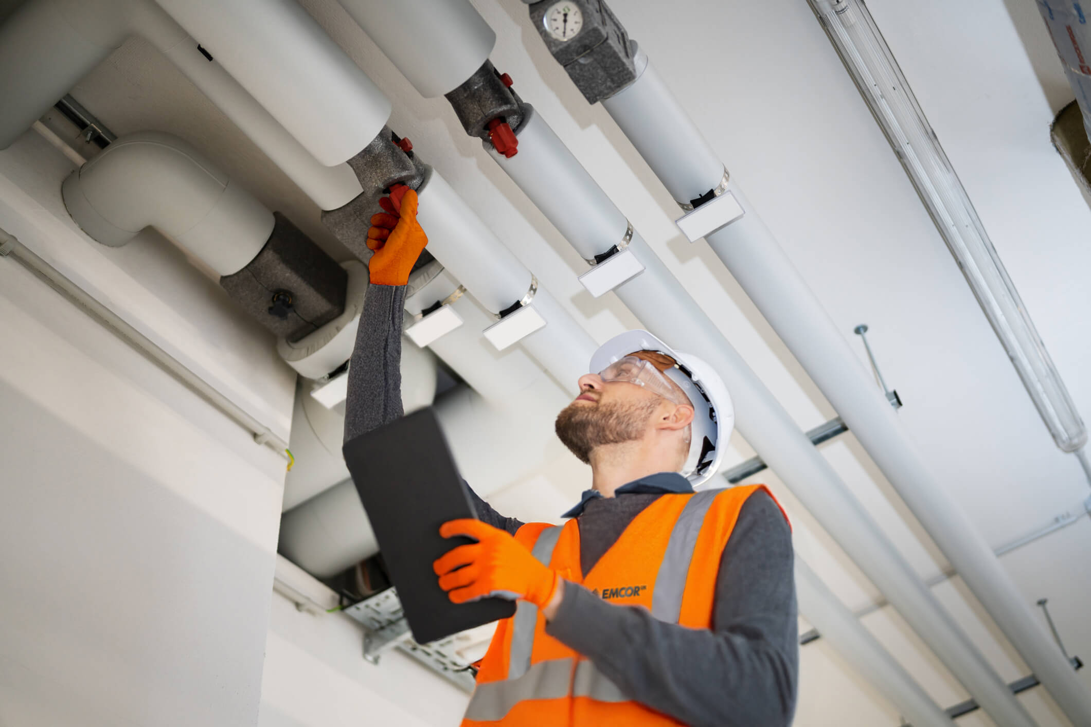 Male water engineer inspecting pipes wearing orange high vis and gloves with hard hat and glasses. 