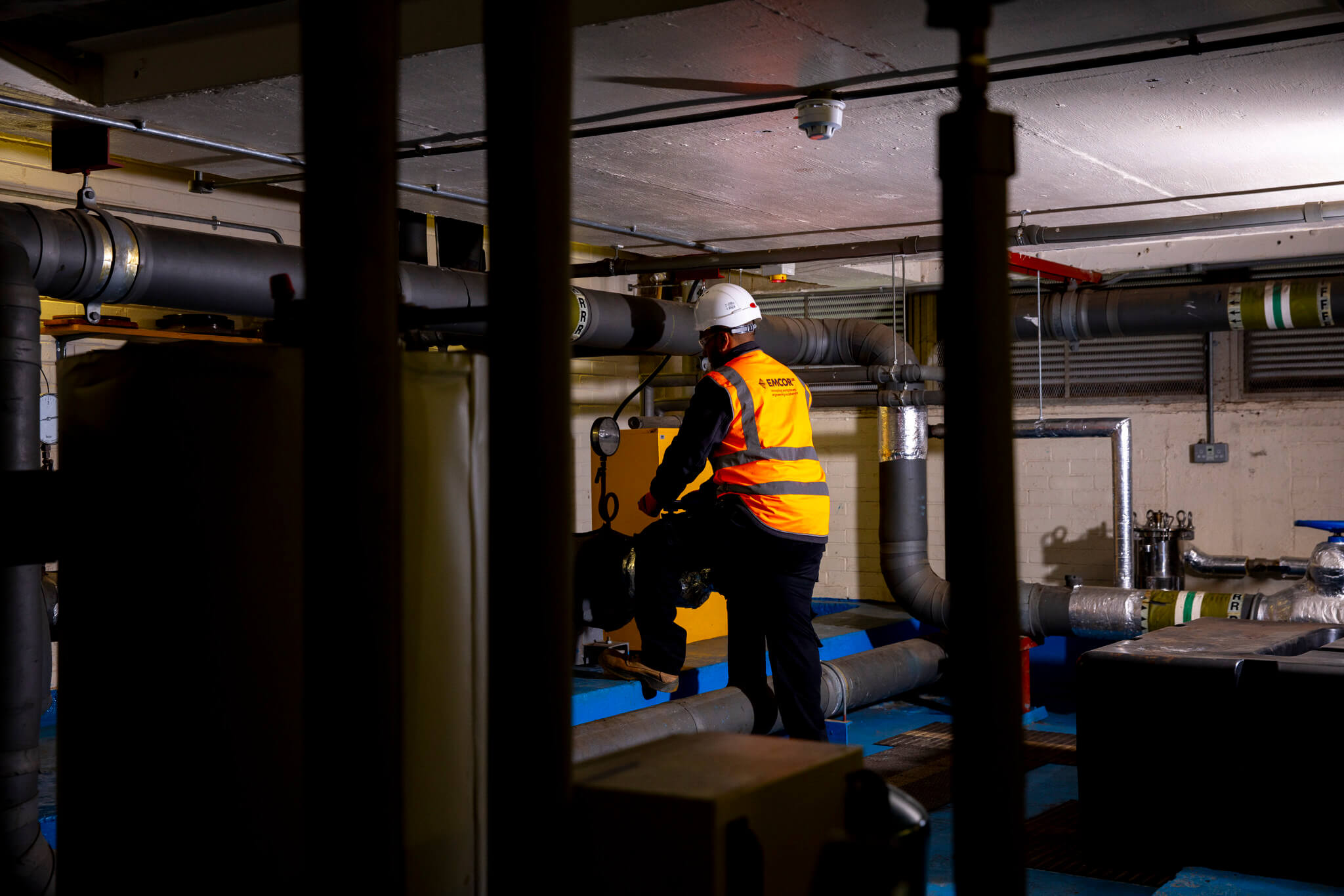 Male engineer wearing orange and black PPE fixing water pipe in a boiler room.