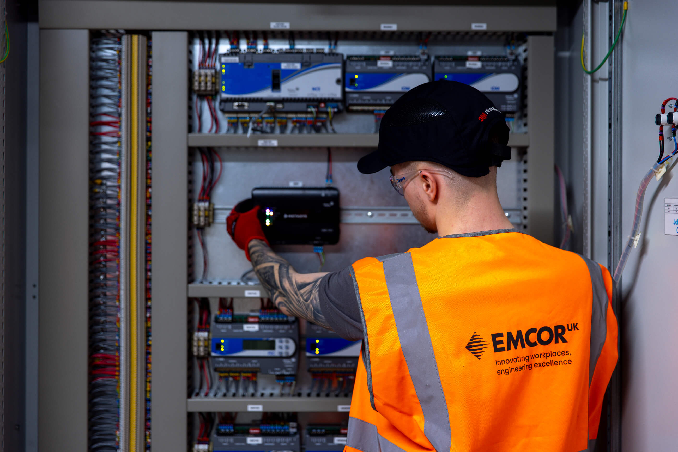 male electrician wearing orange EMCOR UK high vis and hard hat working in an electrical cupboard. 