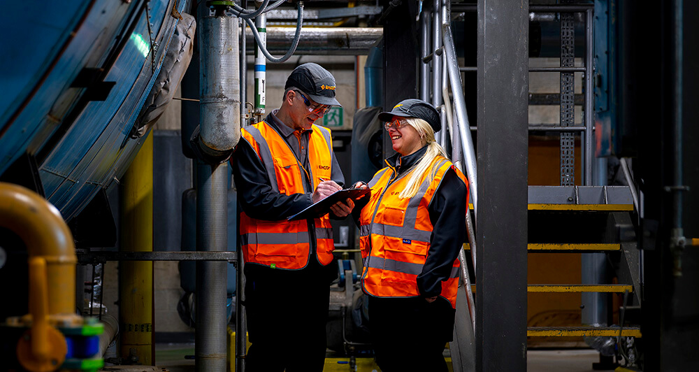 Two EMCOR UK employees wearing orange safety vests at a facility reviewing a clipboard.