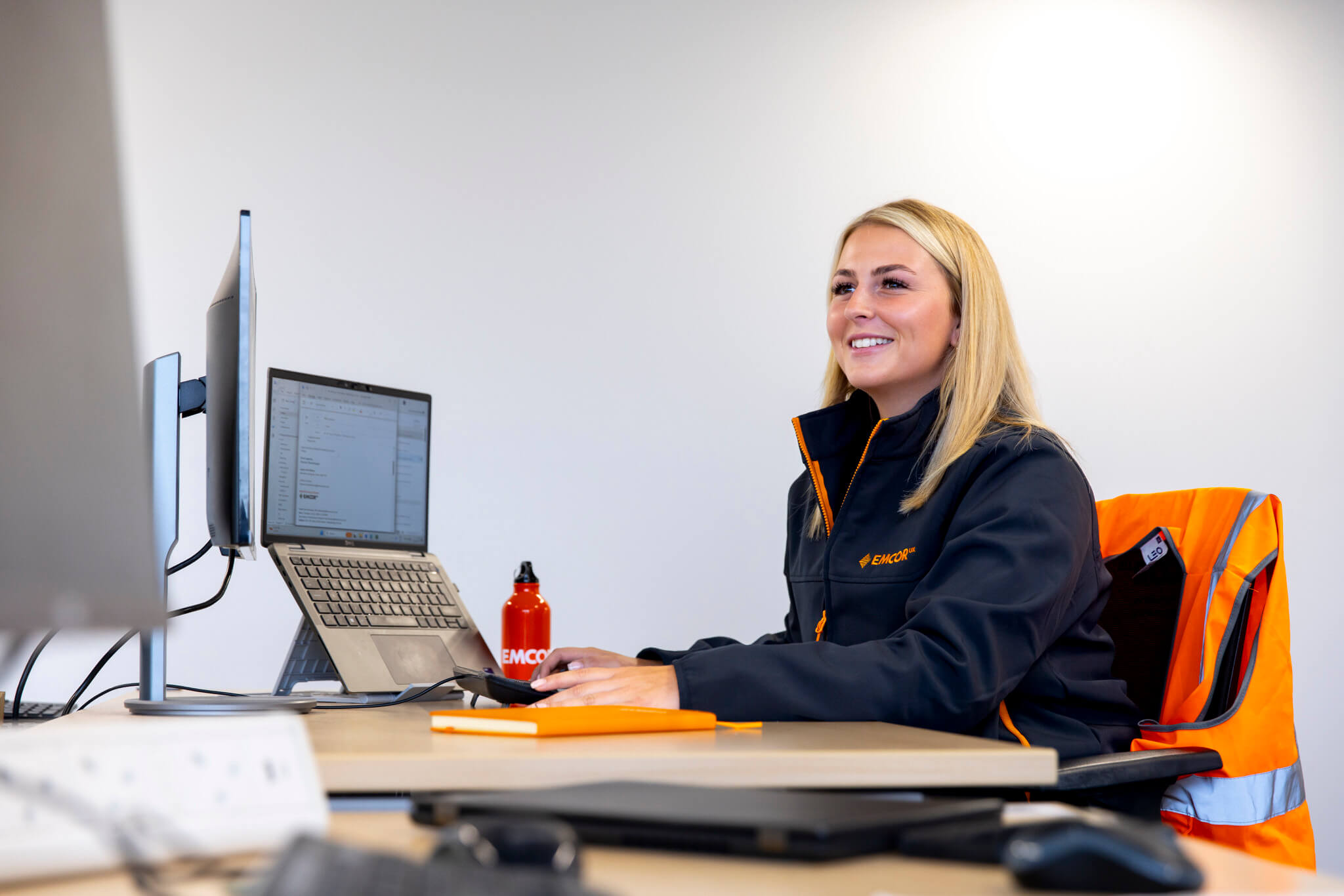 Female worker wearing EMCOR UK uniform sat at a desktop with a laptop and screen.