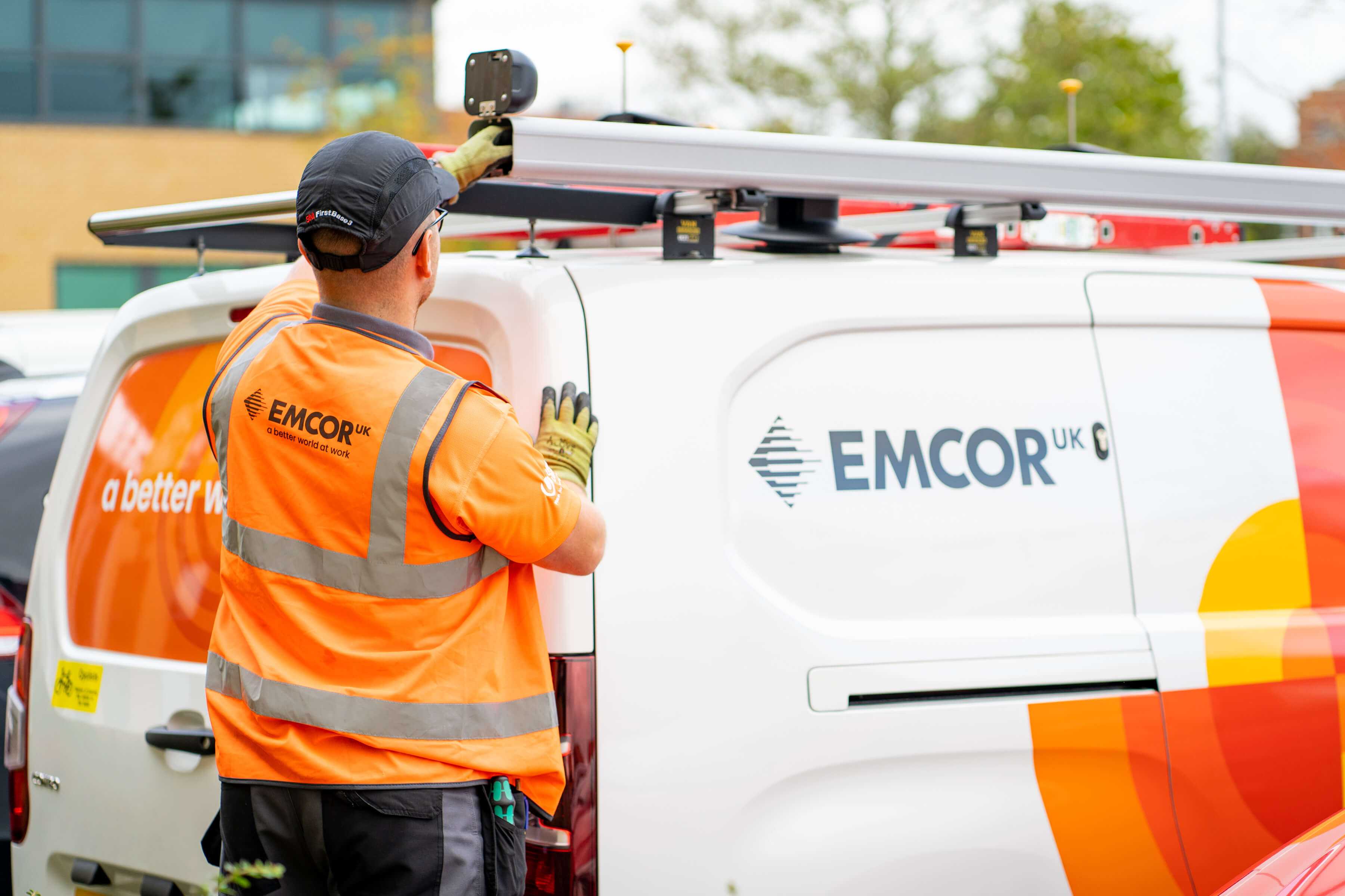 Engineer wearing orange high vis and black cap loading EMCOR UK branded van.