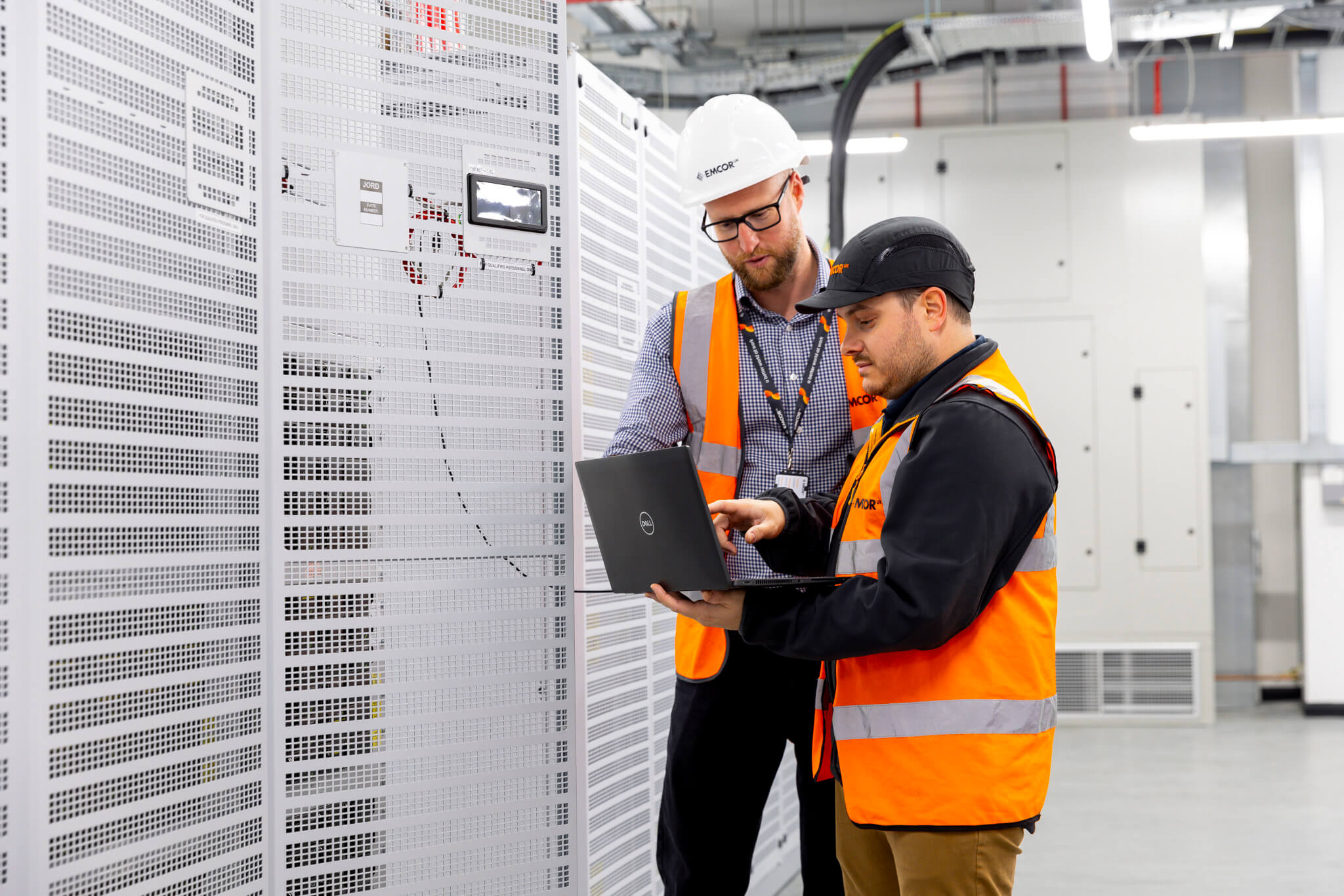 Two male engineers wearing orange high vis and wearing hard helmets standing collecting data on a laptop. 