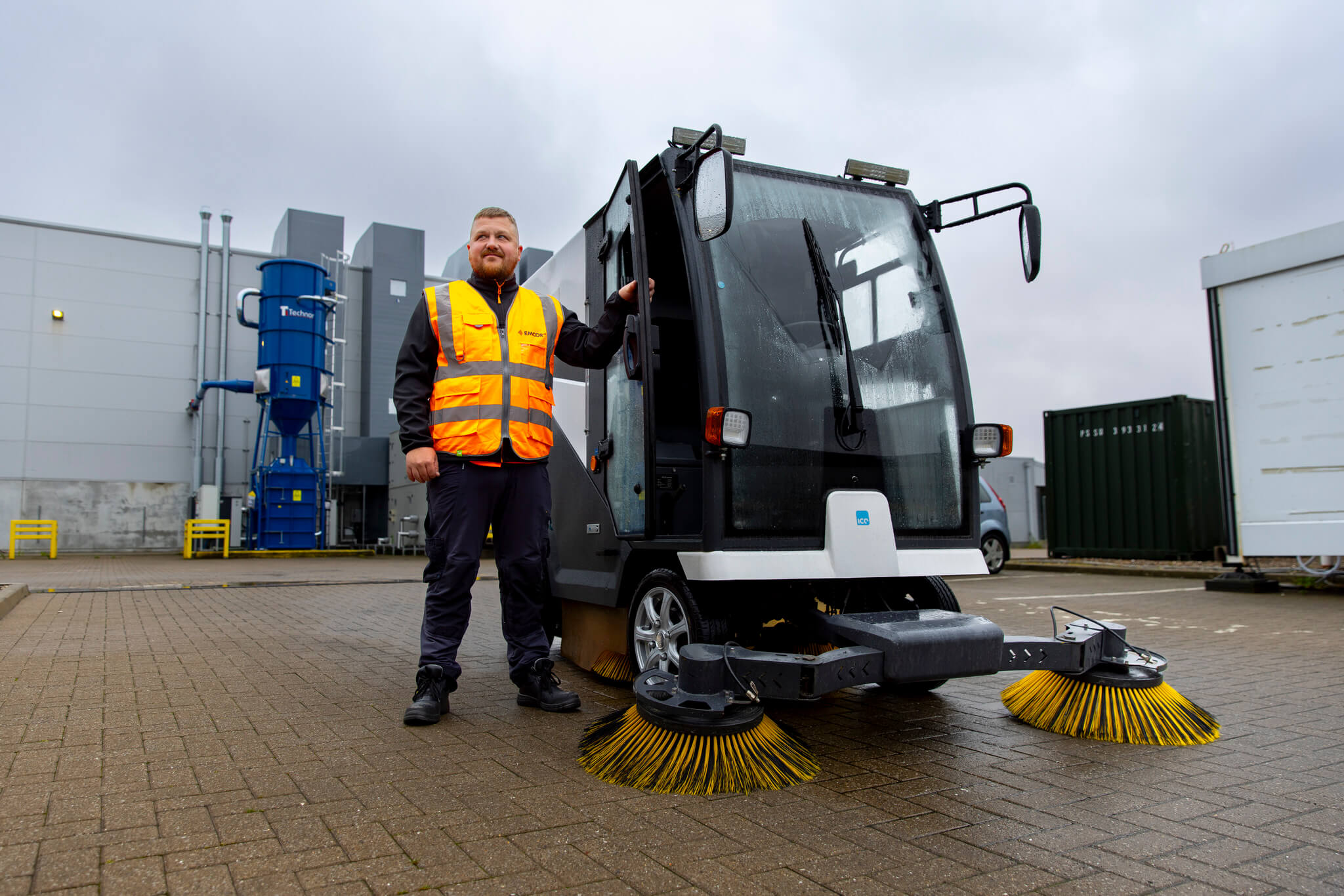 EMCOR UK colleague stood outside next to street sweeper, wearing orange high vis on a cloudy day.