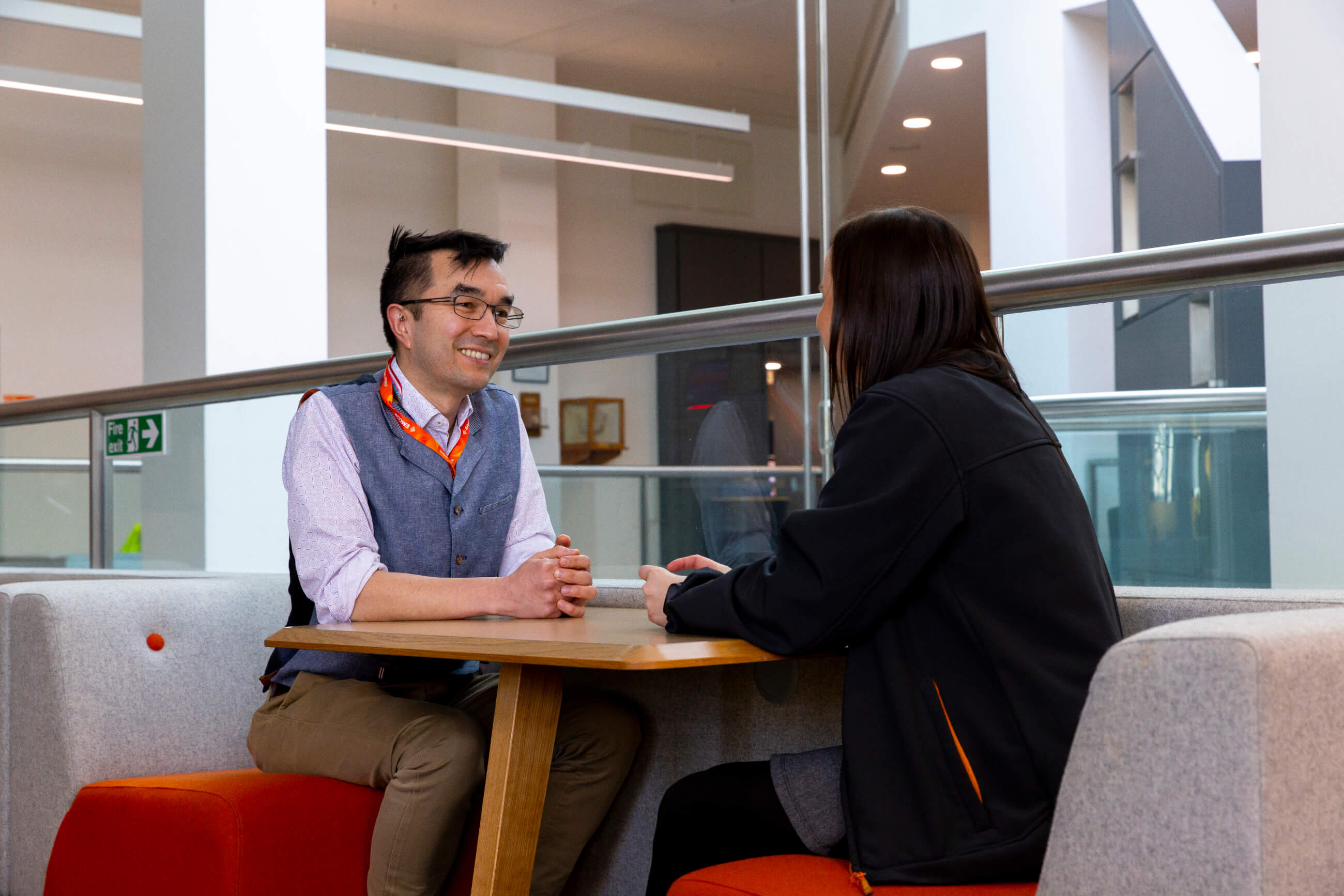 Six colleagues sat in seating area in office. All wearing orange lanyards wearing formal wear. Staircase in the background. 