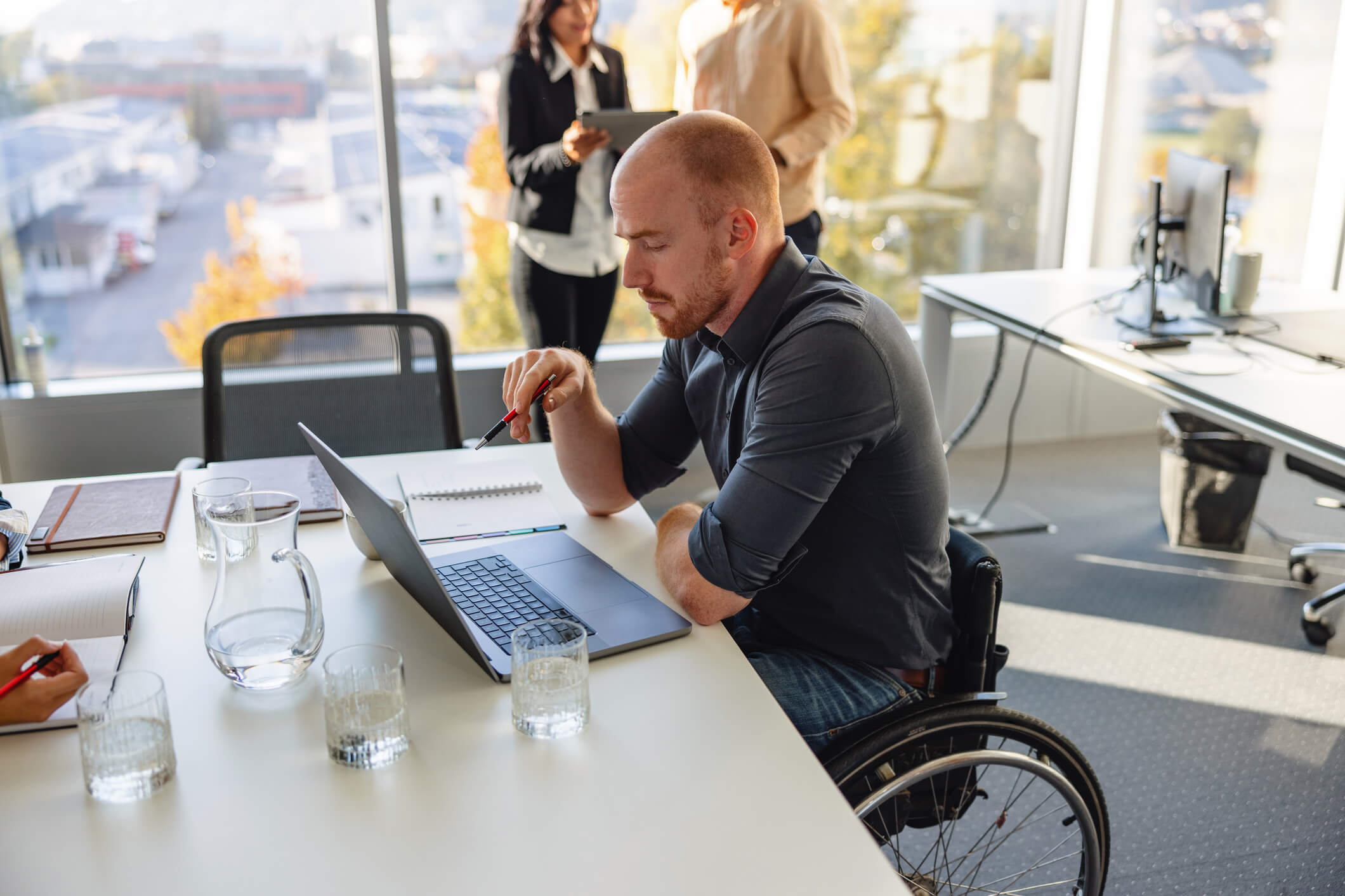 Man in a wheelchair sat at a desk in office environment with laptop and water on the table. 