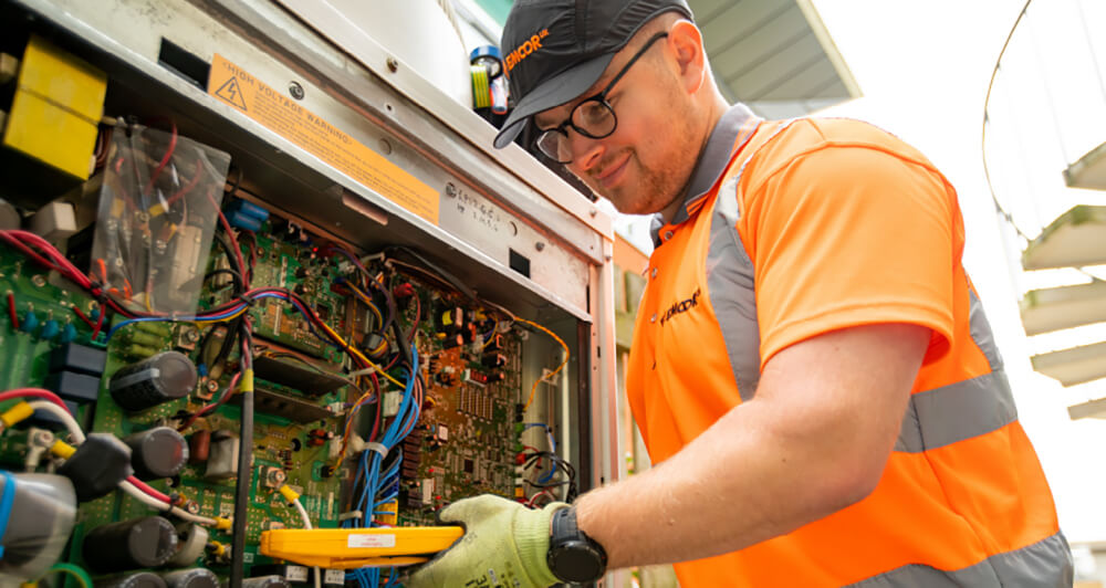 EMCOR UK employee wearing an orange polo shirt and black hat assessing wiring at a client facility.