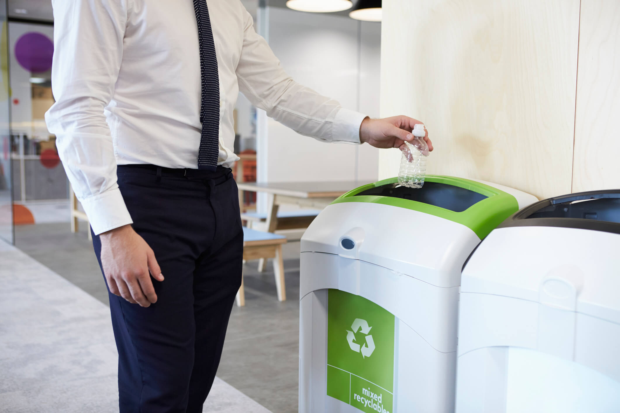 Man wearing white shirt and tie placing a bottle in recycling bin in an office.