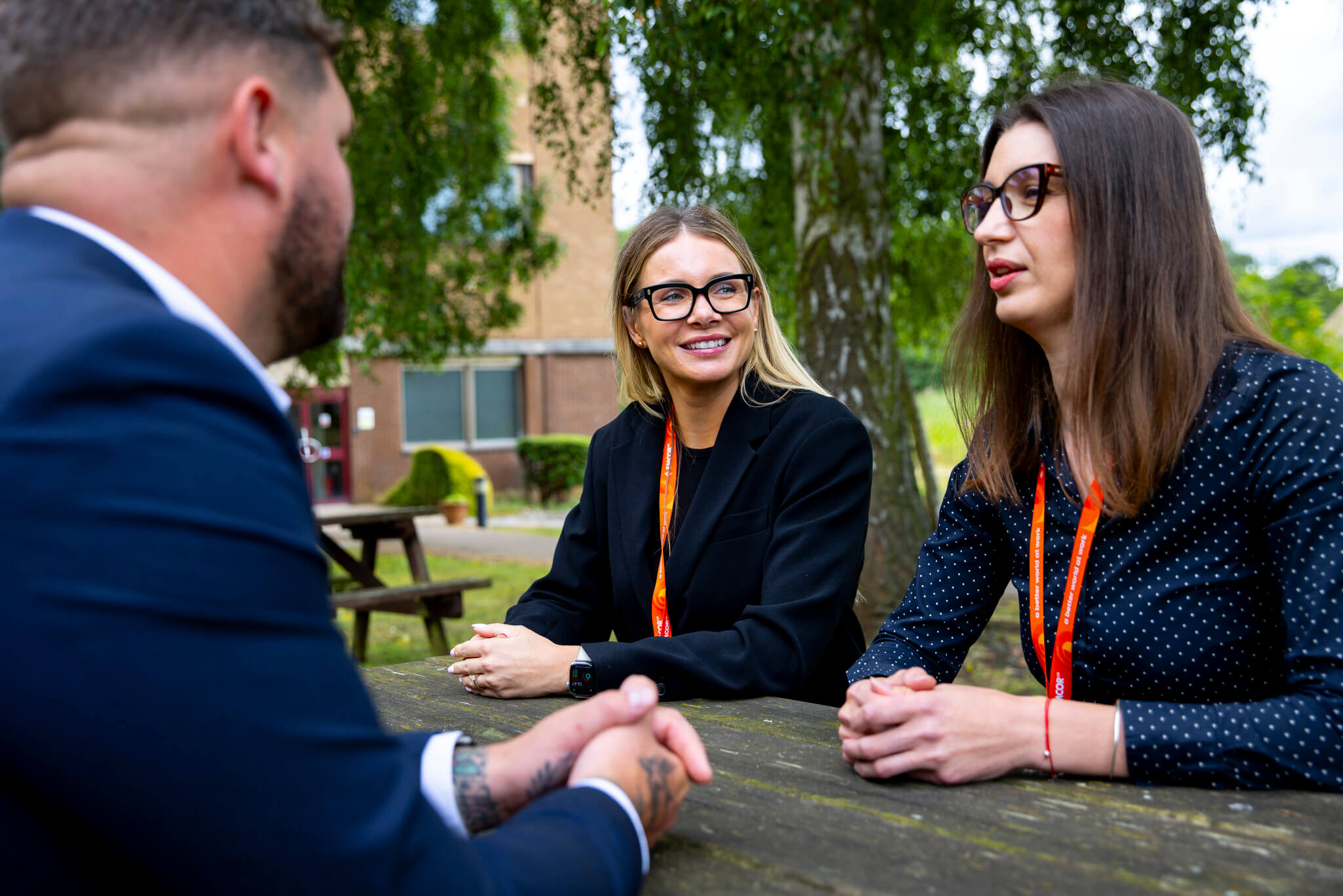 Three colleagues talking while sat out on a bench outside. Trees and office building in the background. 