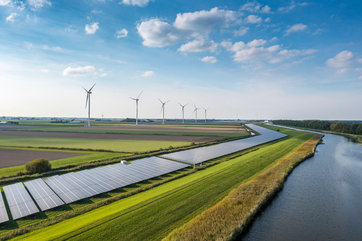 Landscape view wind turbines and solar panels alongside greenery and a river.