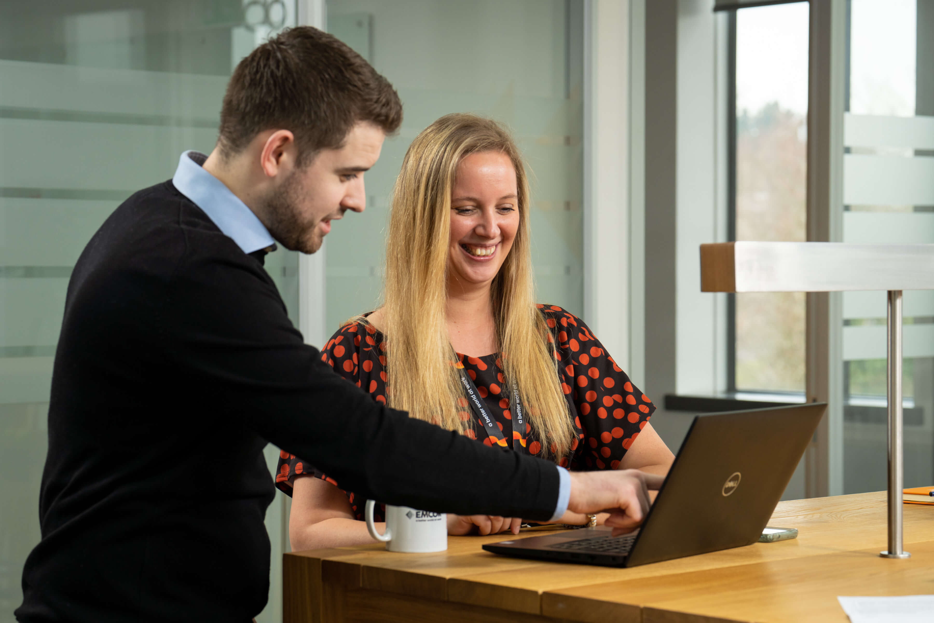 Male and female colleagues working together on laptop. Glass meeting room screens in the background.