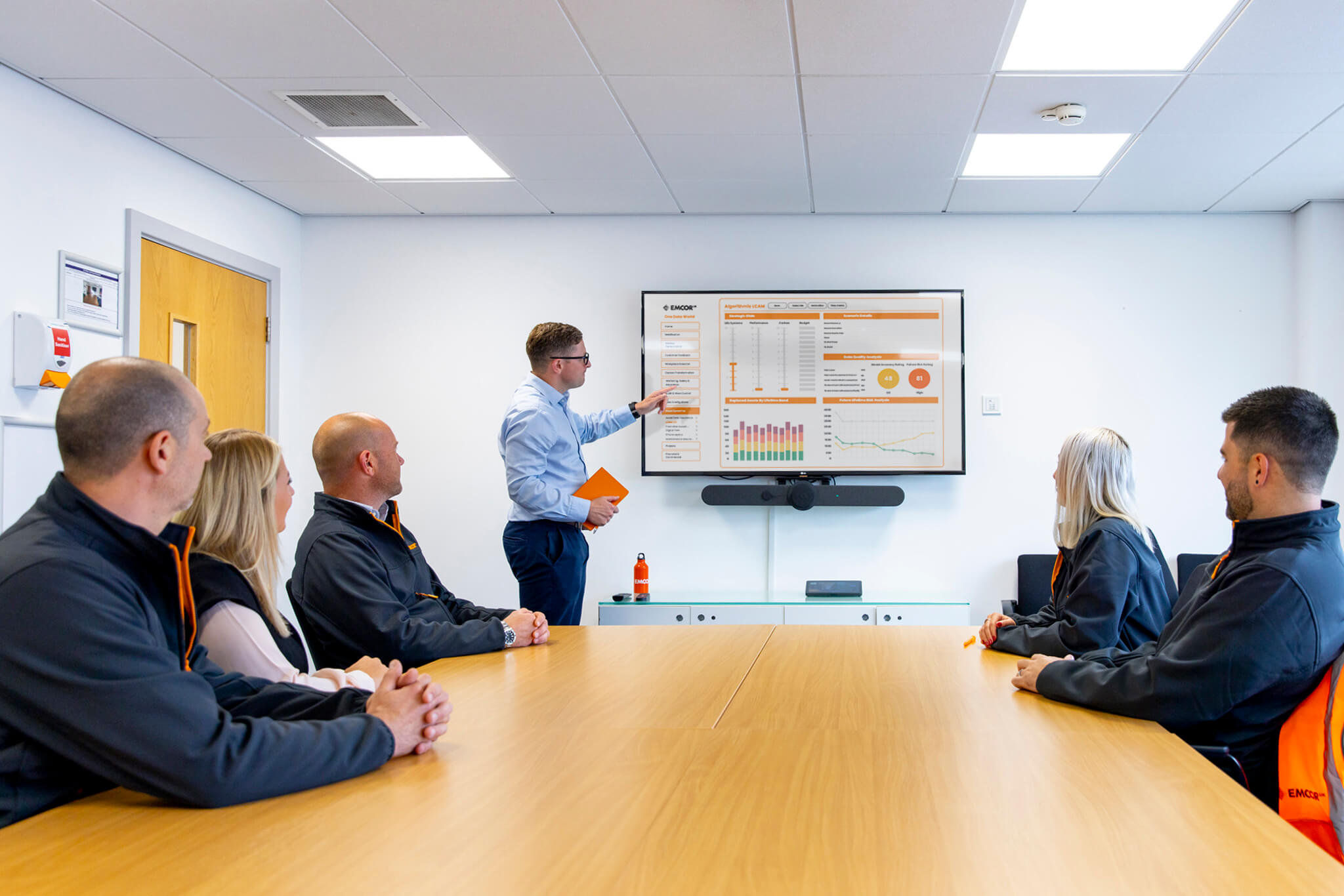 Male colleague presenting to a group of people in large meeting room. Presenting on a large TV screen showing data.