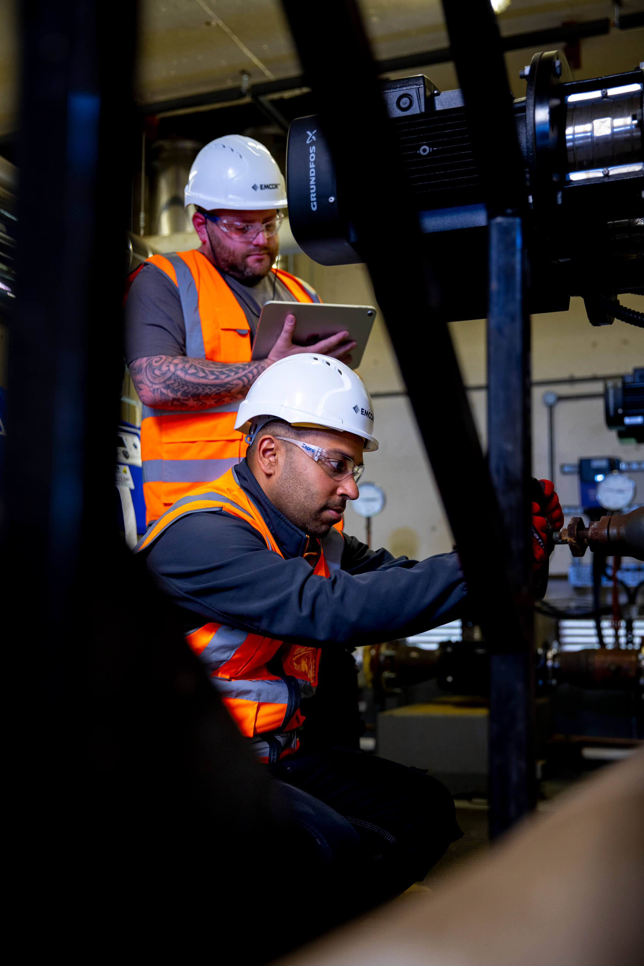 Two male engineers in a boiler room. Male in the back is holding an iPad while his colleague is turning a water valve