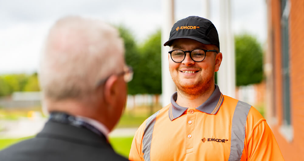 EMCOR UK employee wearing an orange shirt and black hat having a conversation with another man.