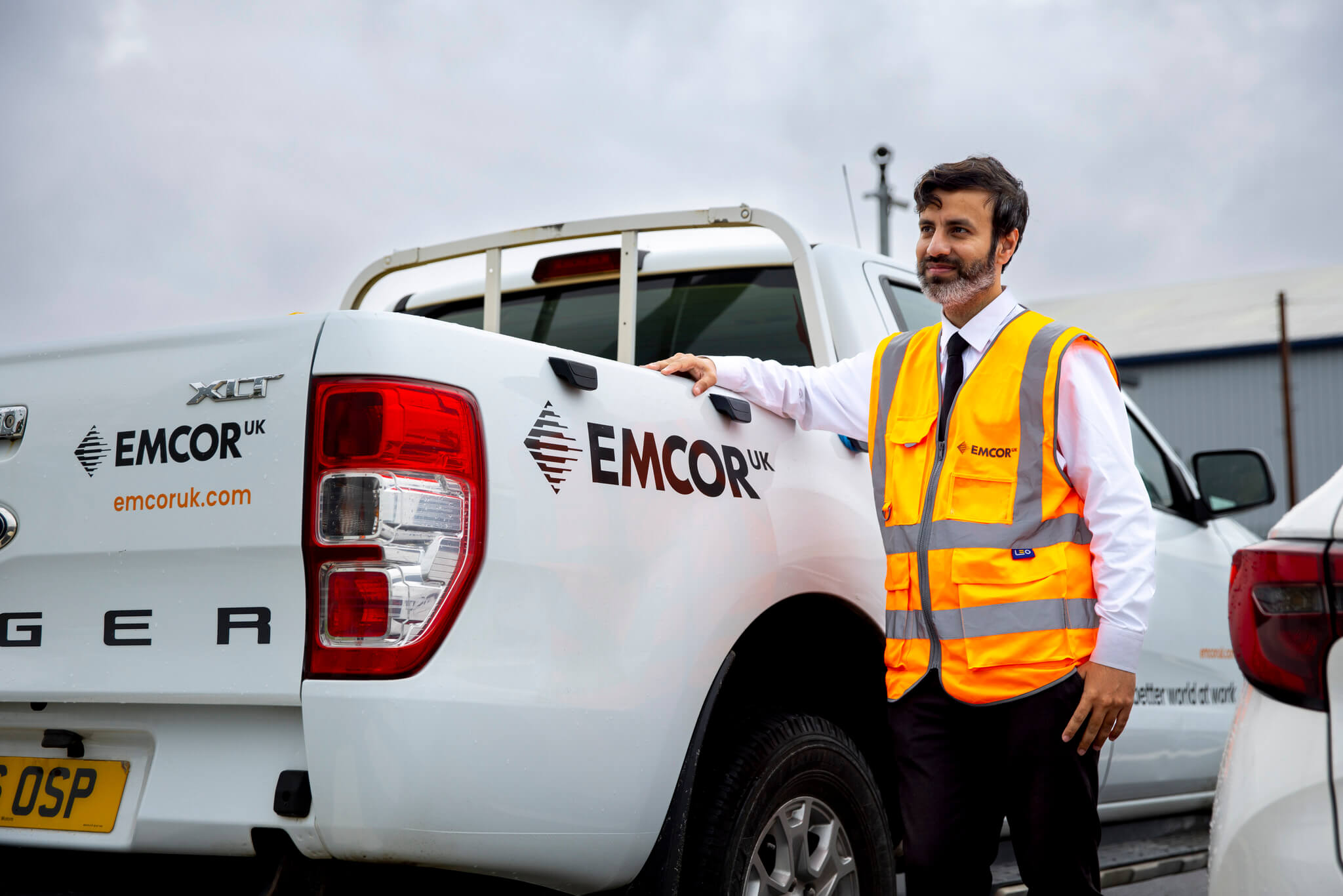 Security guard wearing orange high vis stood next to EMCOR UK branded vehicle.