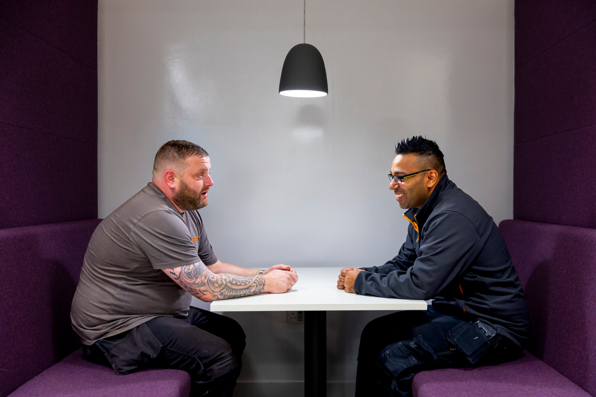 Two male colleagues talking opposite each other in an office booth with purple chairs.