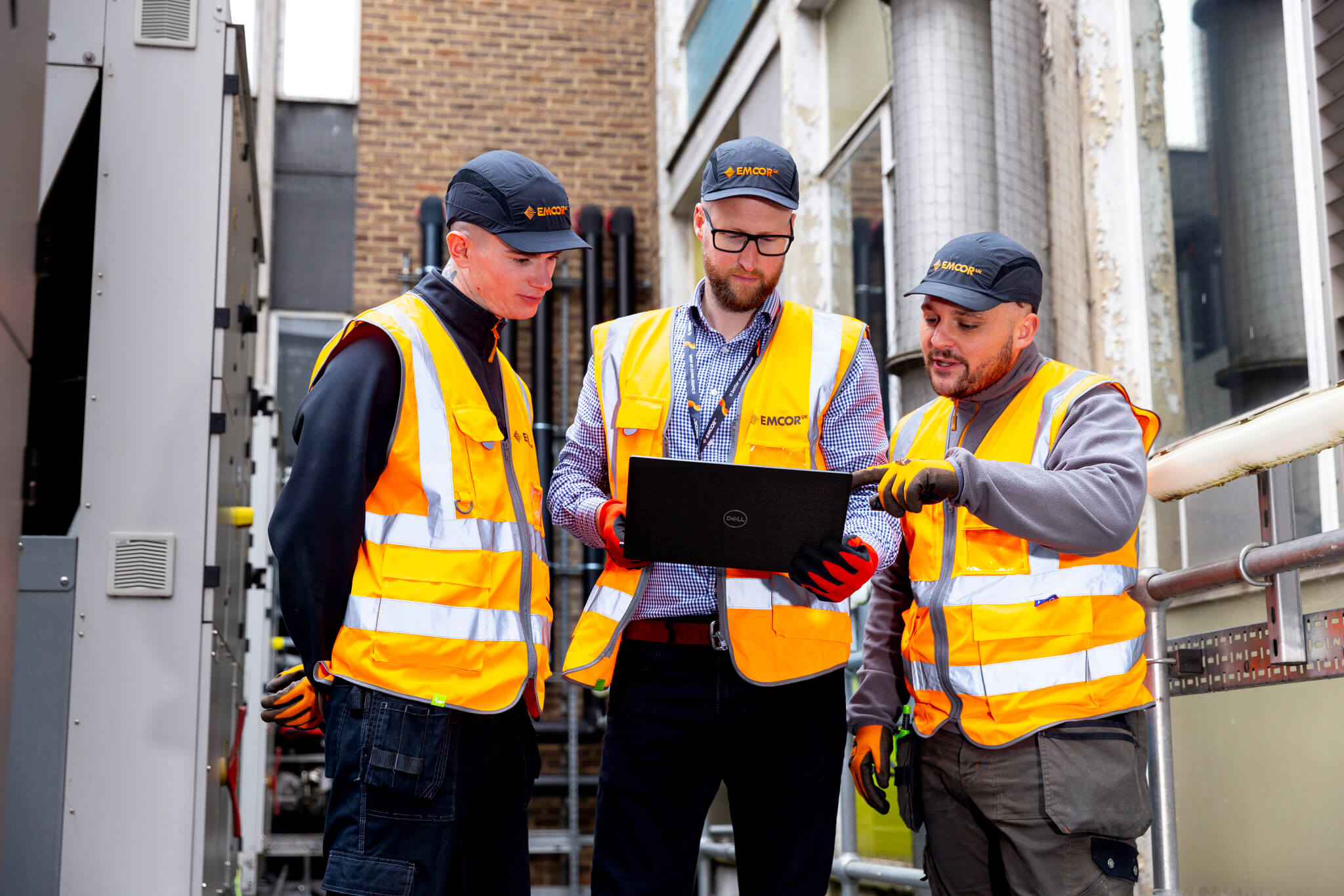 Three male engineers stood outside next to a generator. Person in the middle is holding a laptop,  the others look at it. 