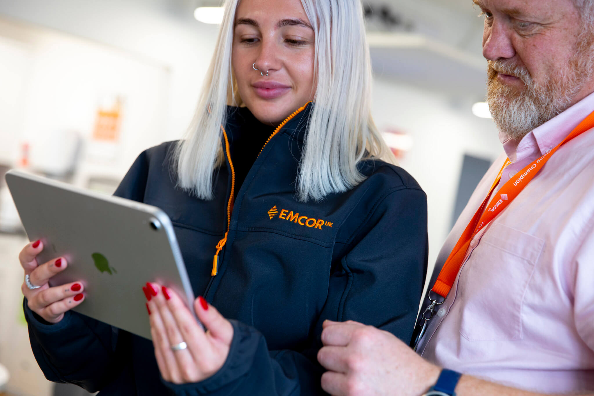 Female colleague in dark grey jacket holding iPad. Male colleague stood slightly behind her looking at the screen.