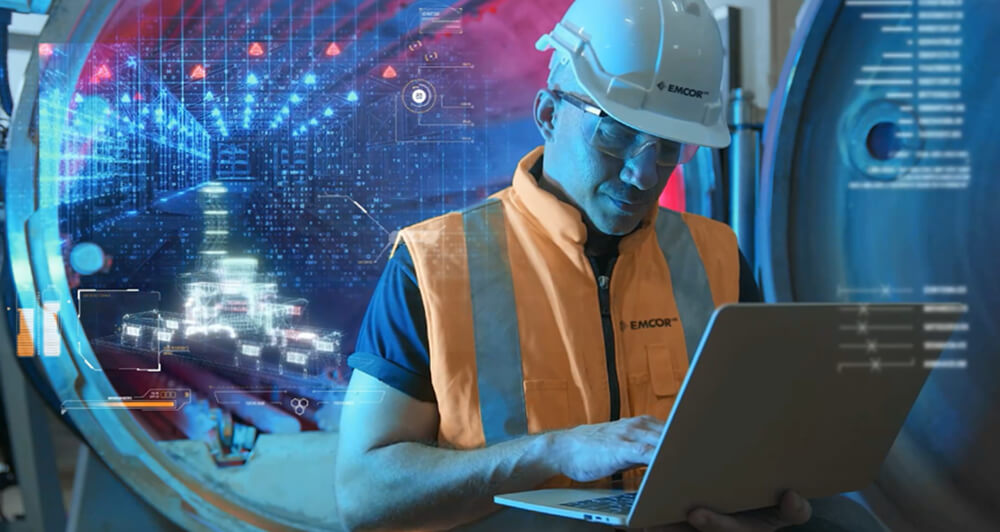 Illustration background of digital data with an EMCOR UK male employee wearing safety gear working on a laptop.