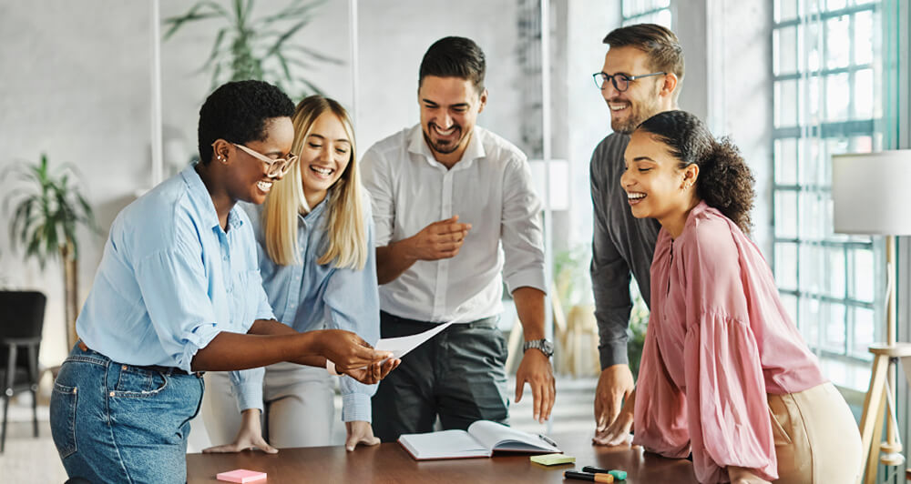 Five coworkers standing around a conference room table discussing documents