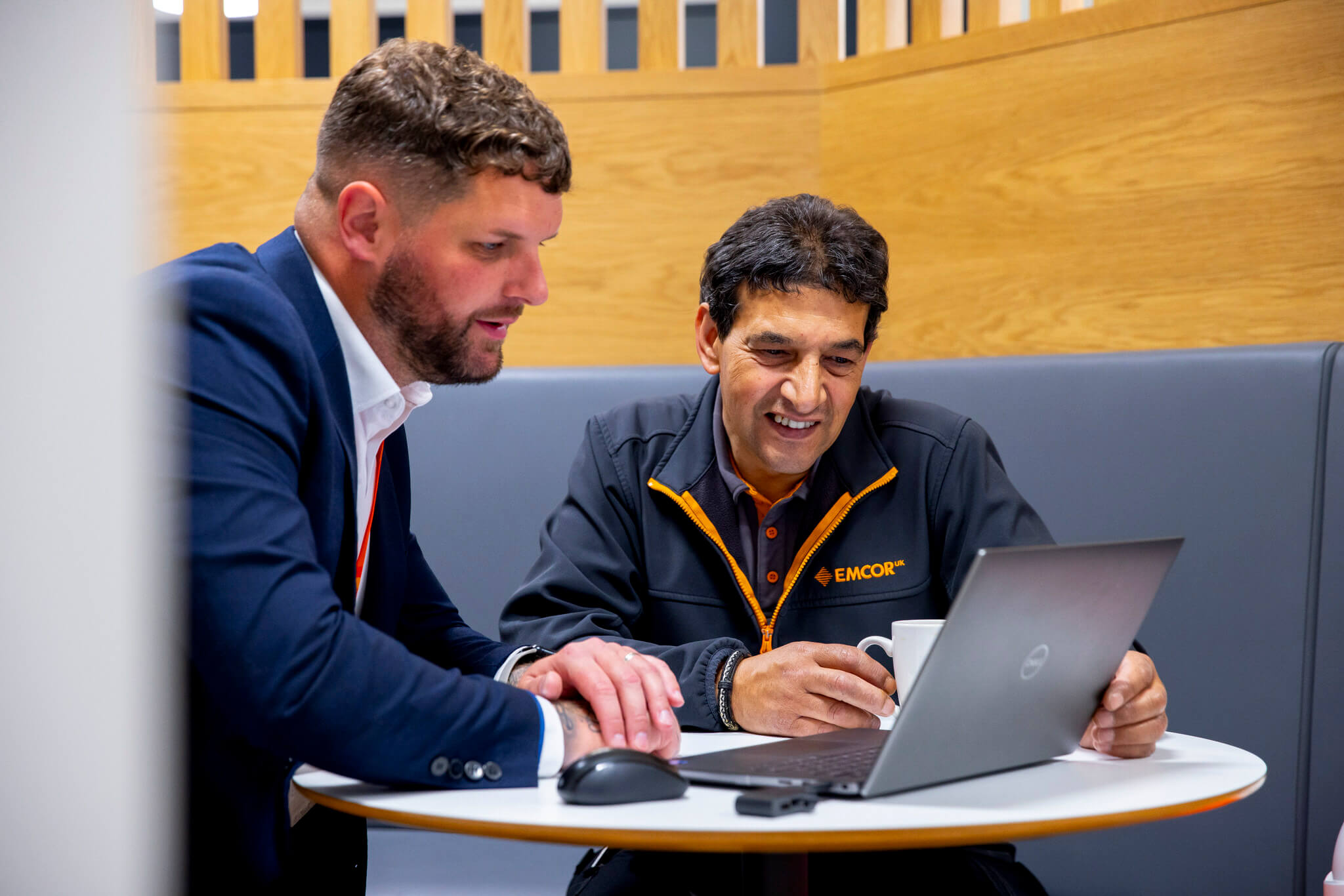 Two male colleagues, sat at a small table, looking at a laptop screen in a hotdesking area. 