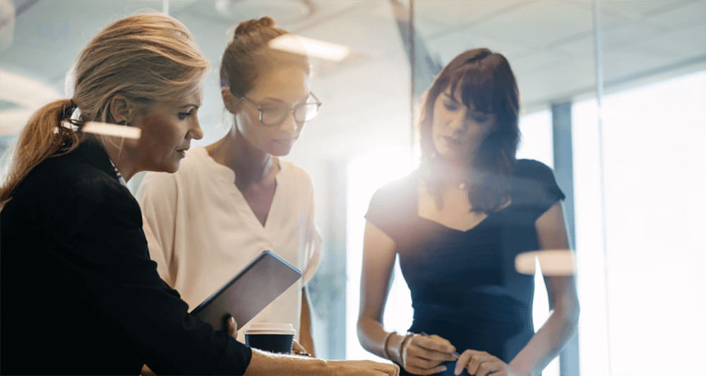 Three female coworkers discussing a project while standing around a table.