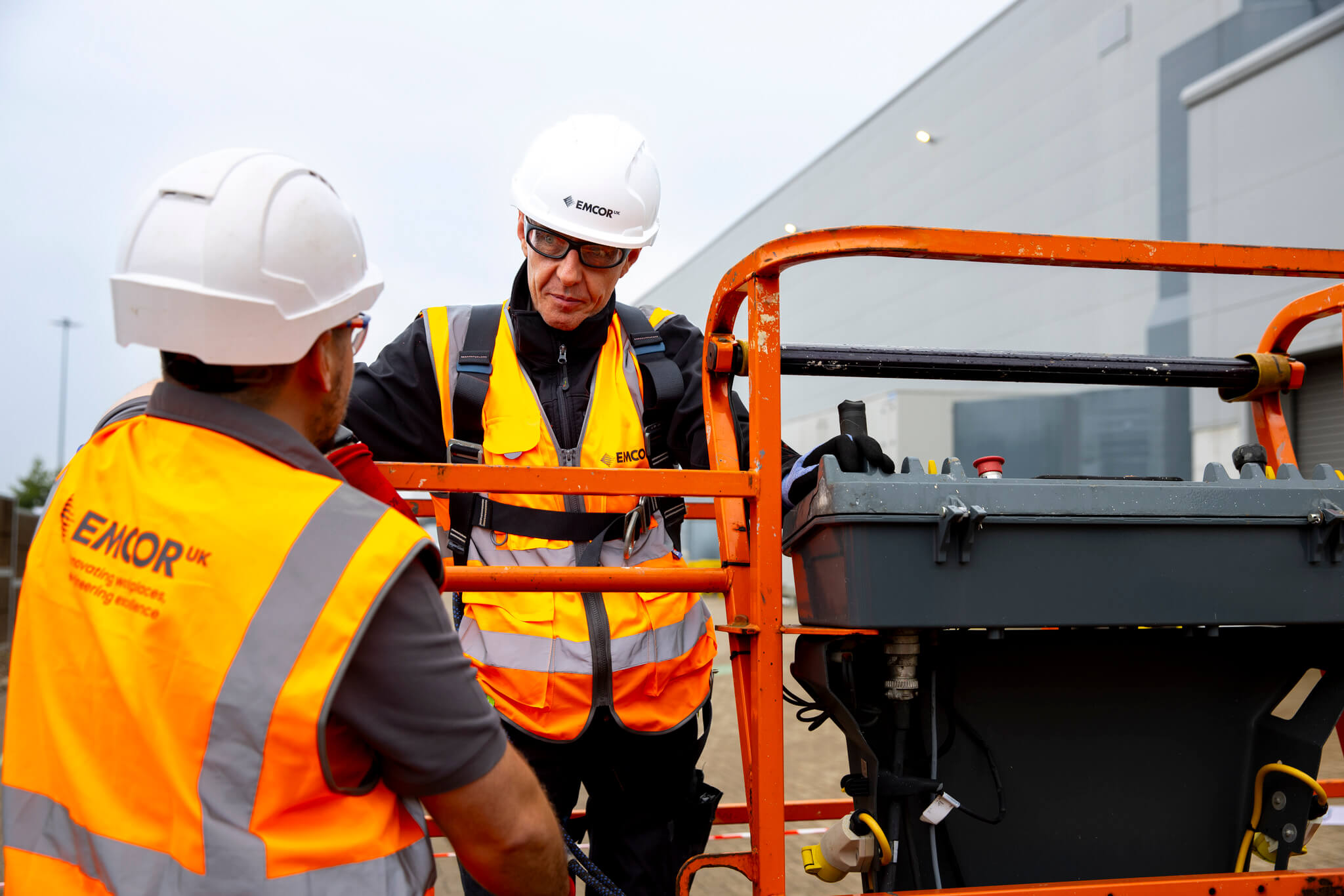 Two engineers talking outside a facility. One is about to operate a scissor lift to conduct building repairs. 