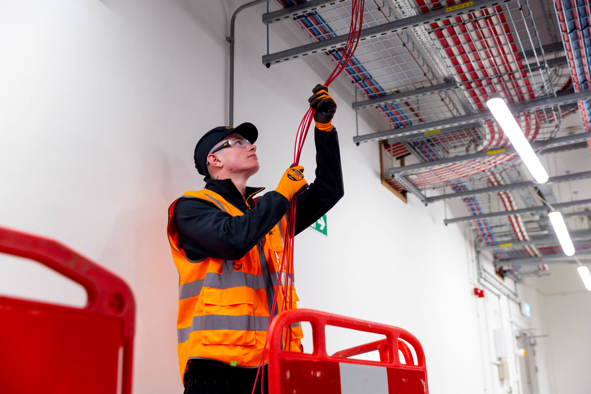 Male engineer holding cables from ceiling. Wearing an orange high vis, glasses, hard hat and gloves surrounded by red barrier.