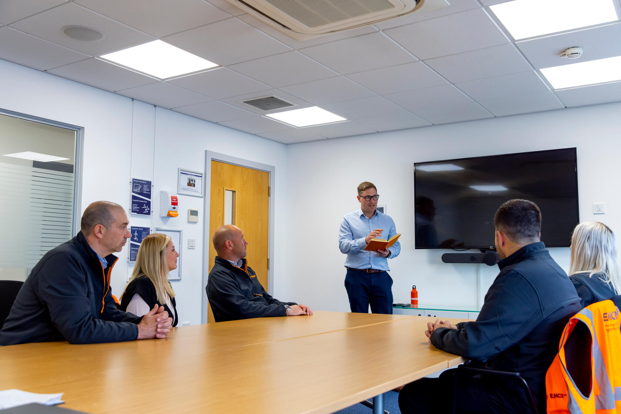 Colleagues sat around a boardroom table having a meeting, with man presenting at the front of the room.