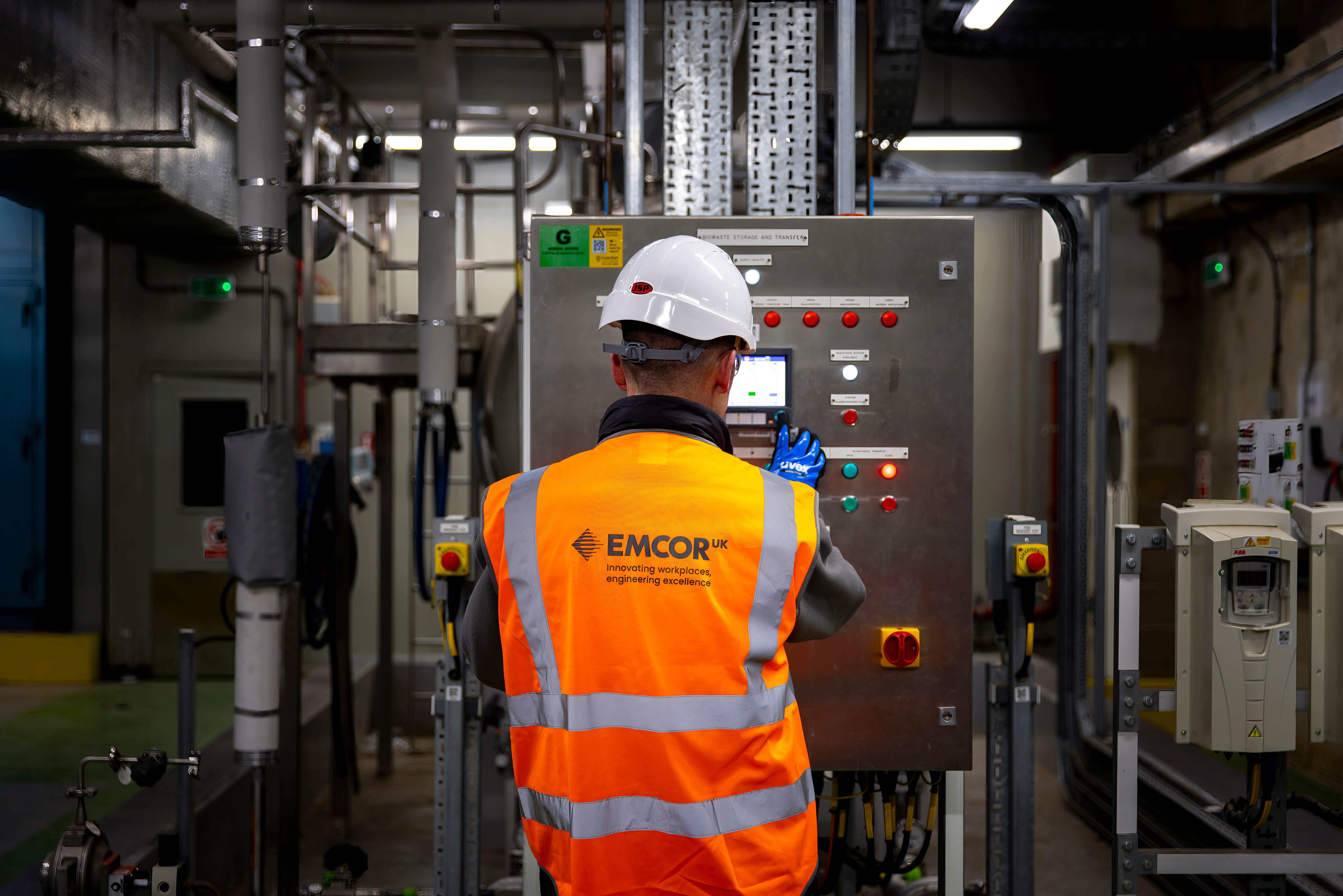 Male engineer wearing orange high vis and white hard hat stood in front of control panel in a boiler room. 