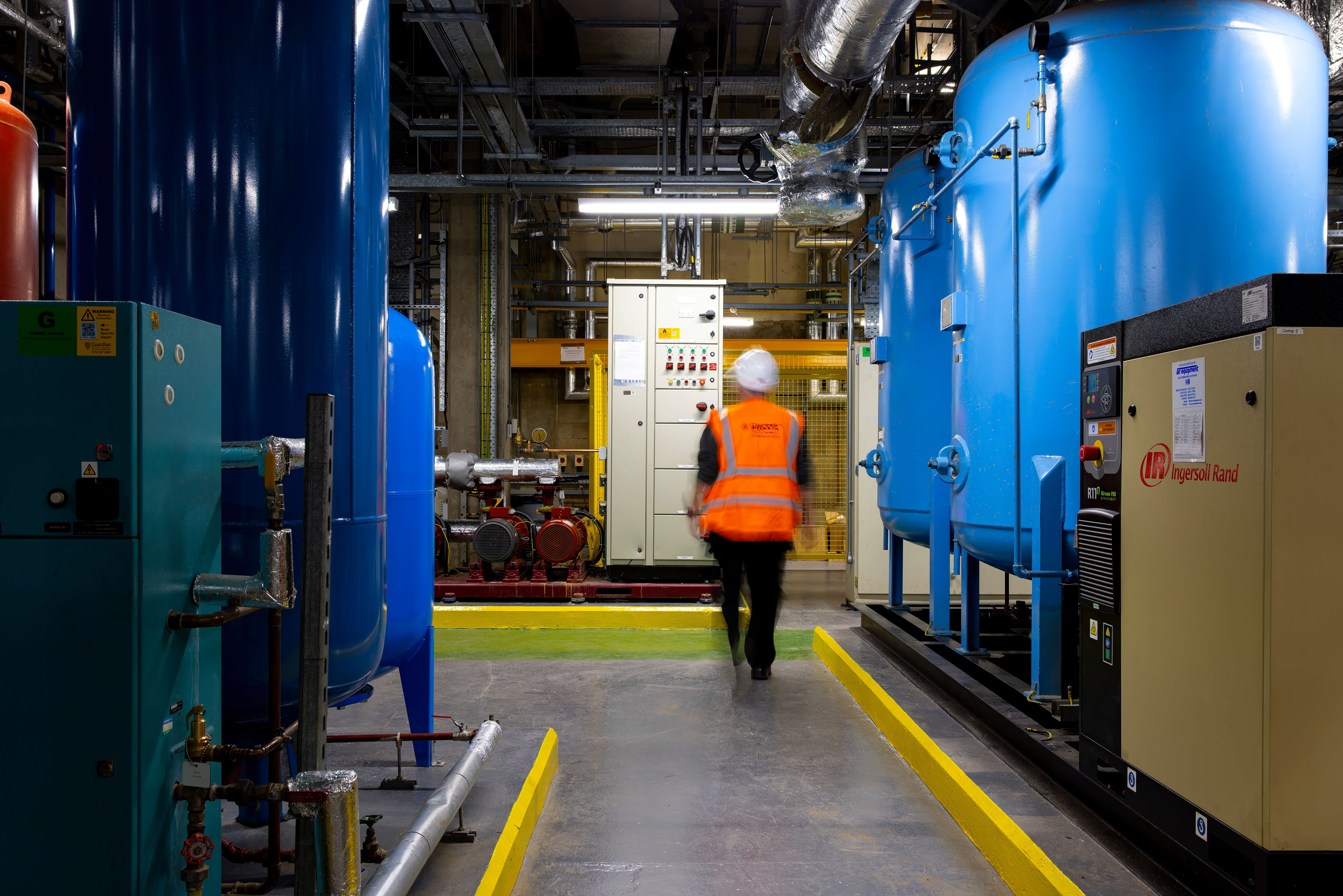 Engineer wearing orange high vis and white hard hat walking through a plant room with mechanical systems on both sides. 