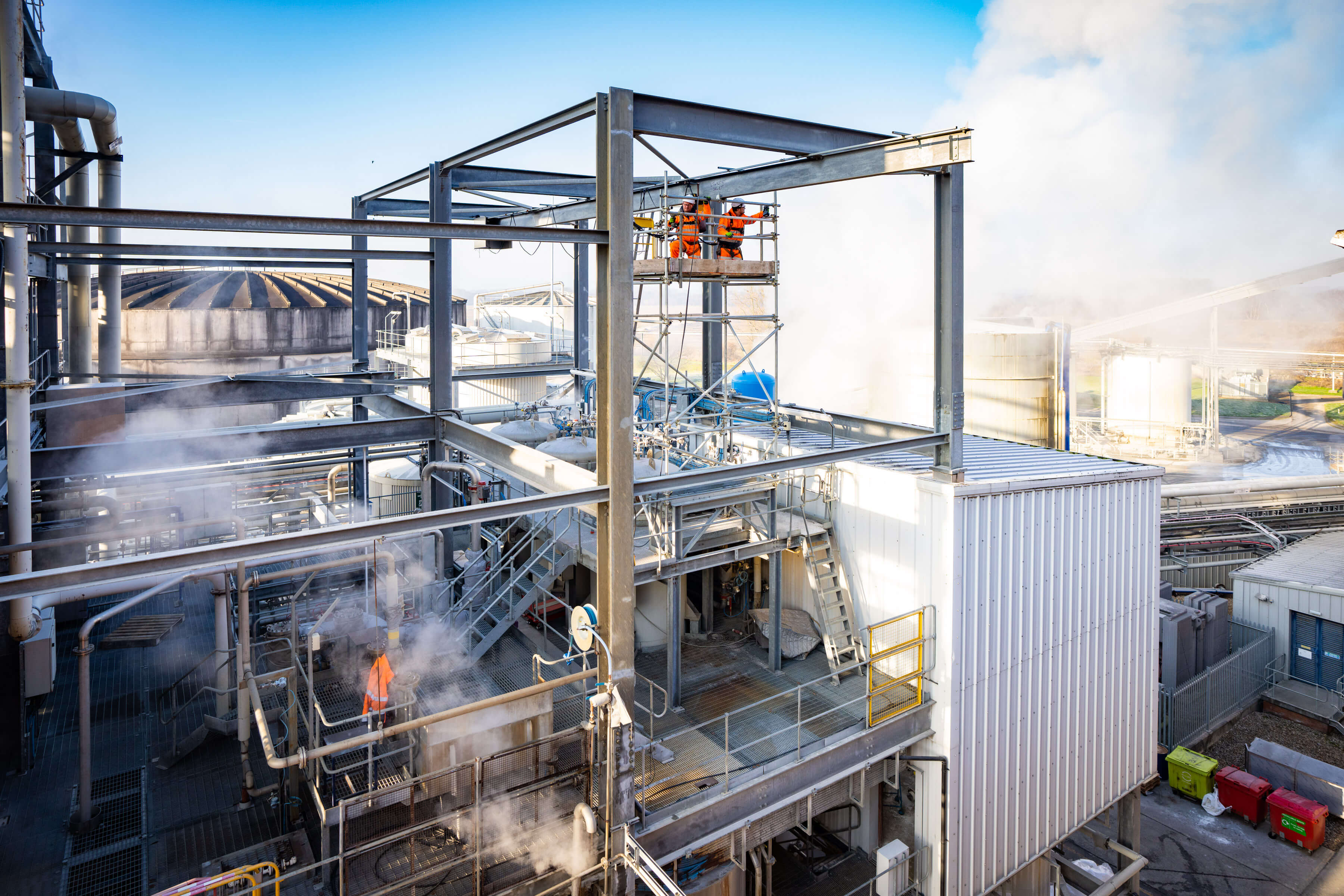 Aerial shot of large industrial facility with steam coming off in multiple areas. Two workers on scaffold platform. 