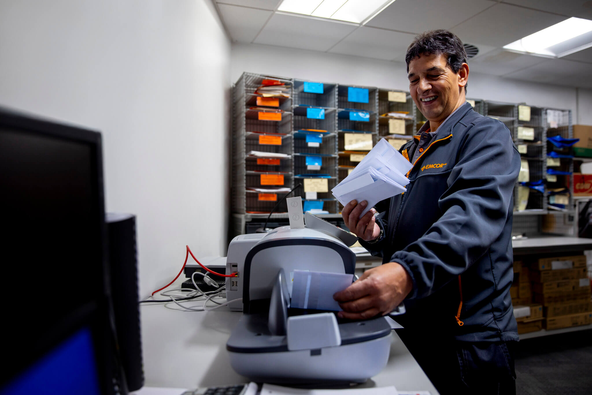 Office worker standing in a post room facing a noticeboard feeding letters through a franking machine.