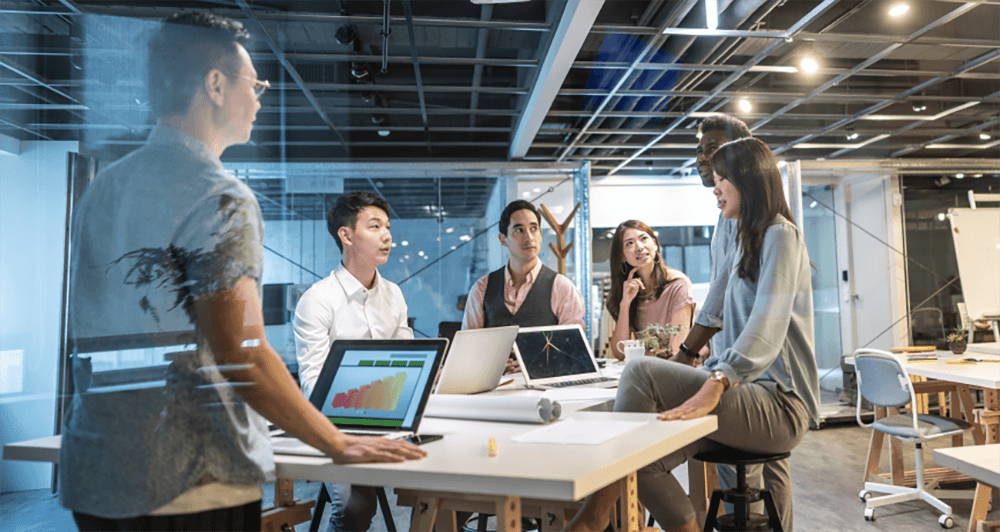 Six business people sitting around a table having a discussion while working on laptops