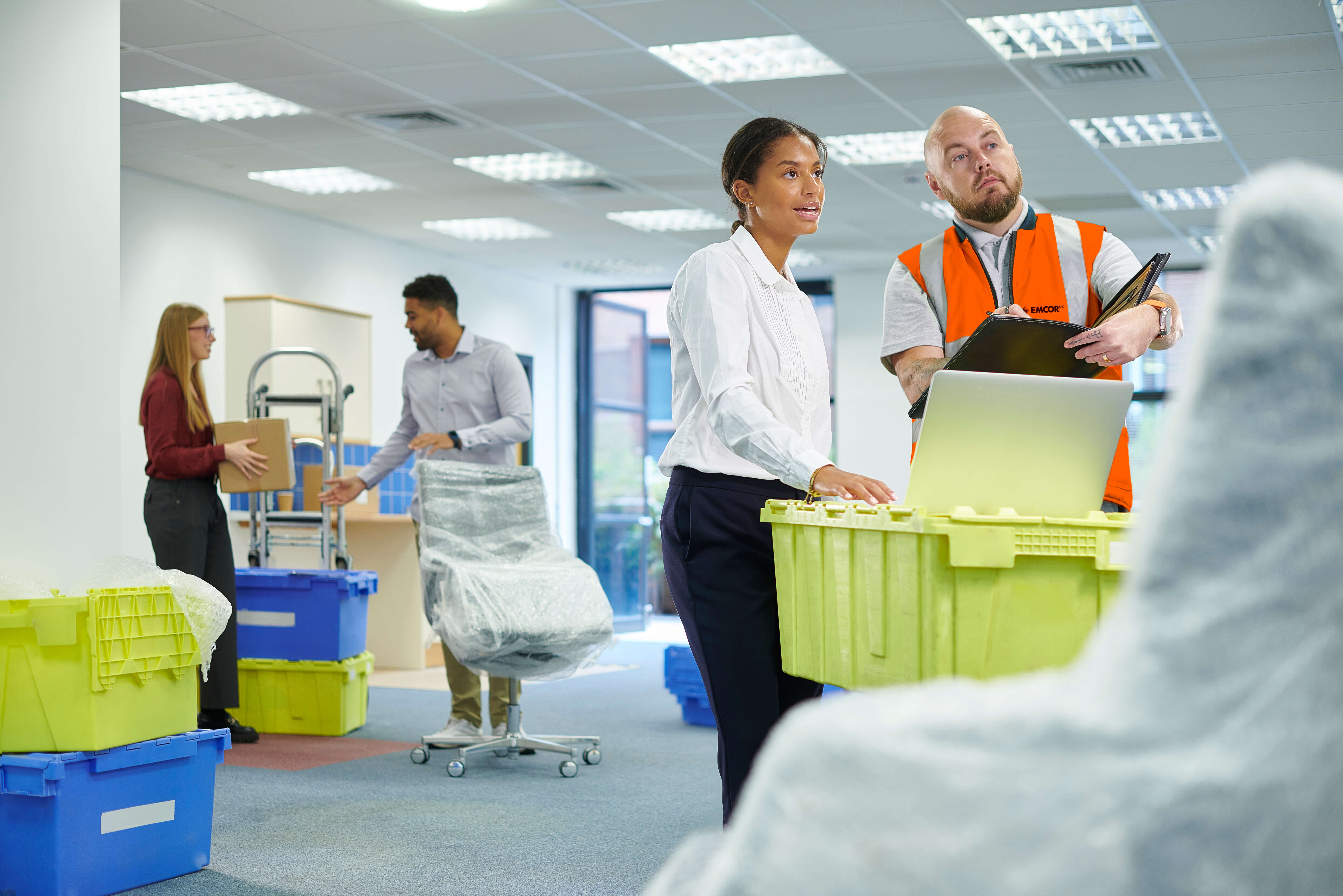 Four people in a large office space. Two looking at plans on laptop. Two in the background unpacking items from boxes. 