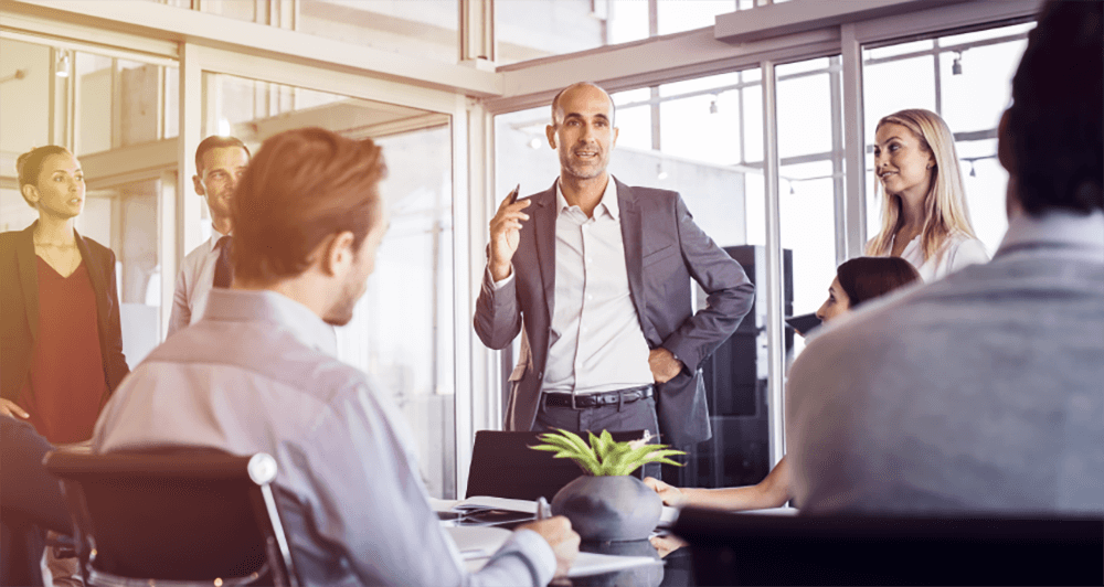 Man wearing a suit talking to a group of coworkers in a corporate setting